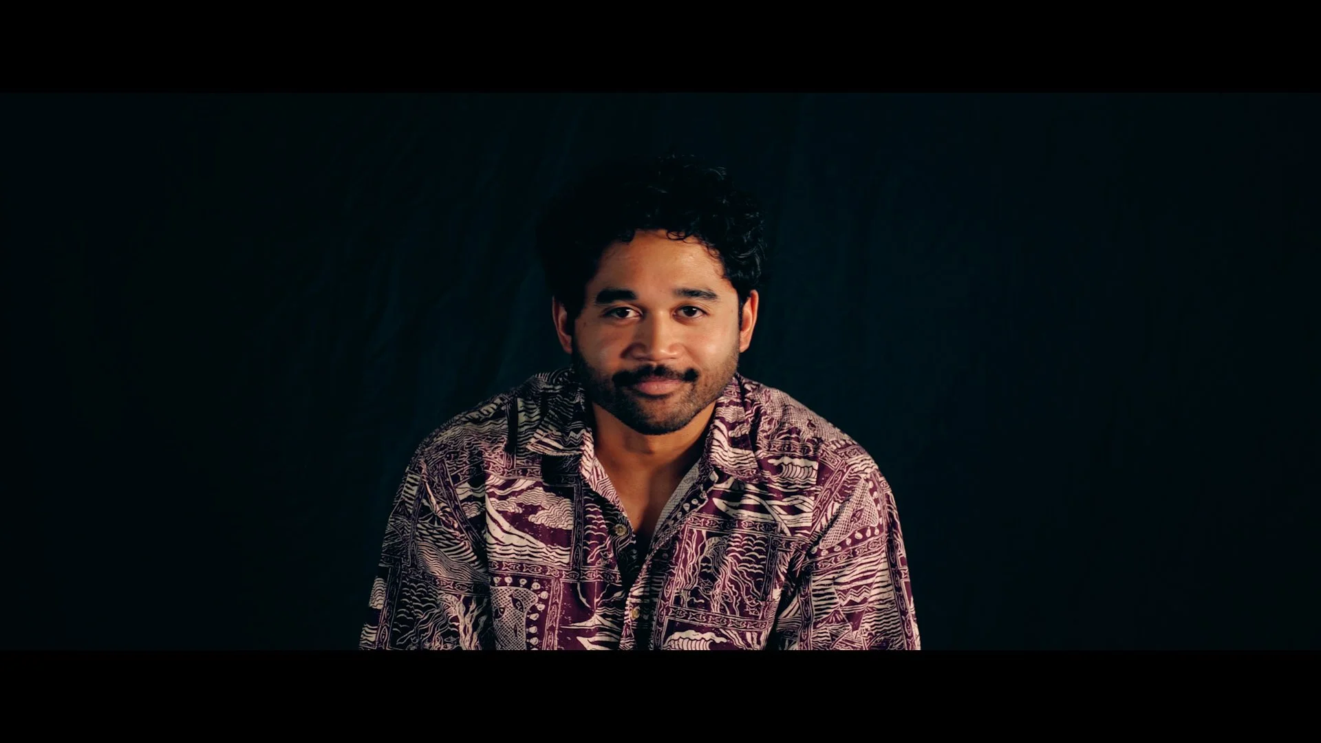 Portrait of a man with dark curly hair and a beard, wearing a patterned shirt, against a dark background.