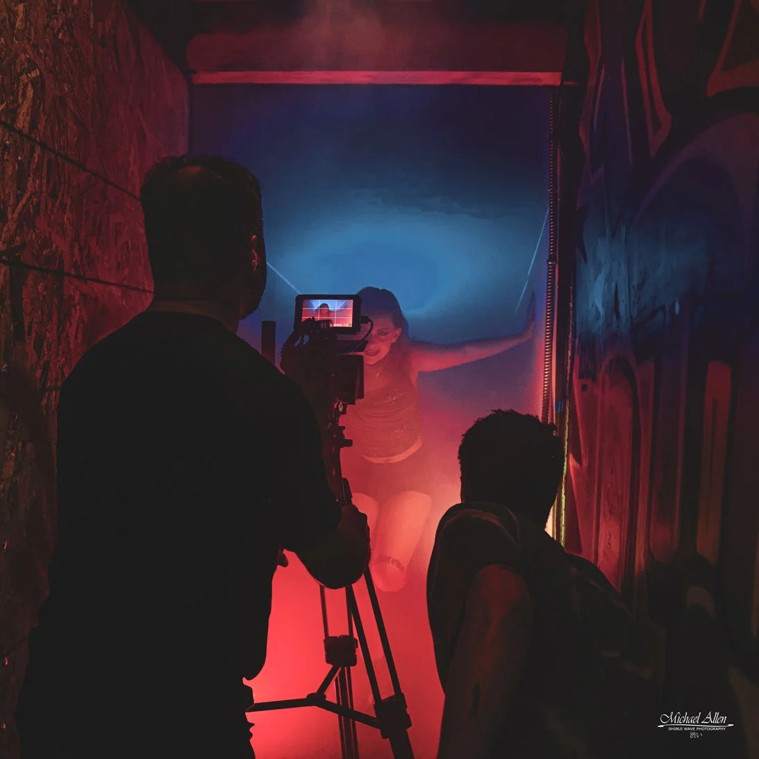 A photographer is taking a picture of a woman posing in a colorful, smoky environment with blue and red lighting, with graffiti on the walls.