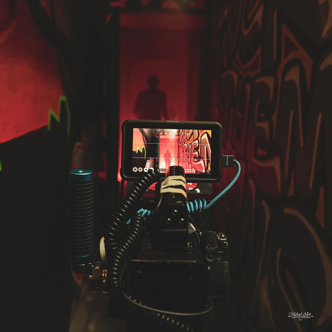 A camera filming a silhouette of a person walking down a narrow, graffiti-covered corridor illuminated with red lighting.