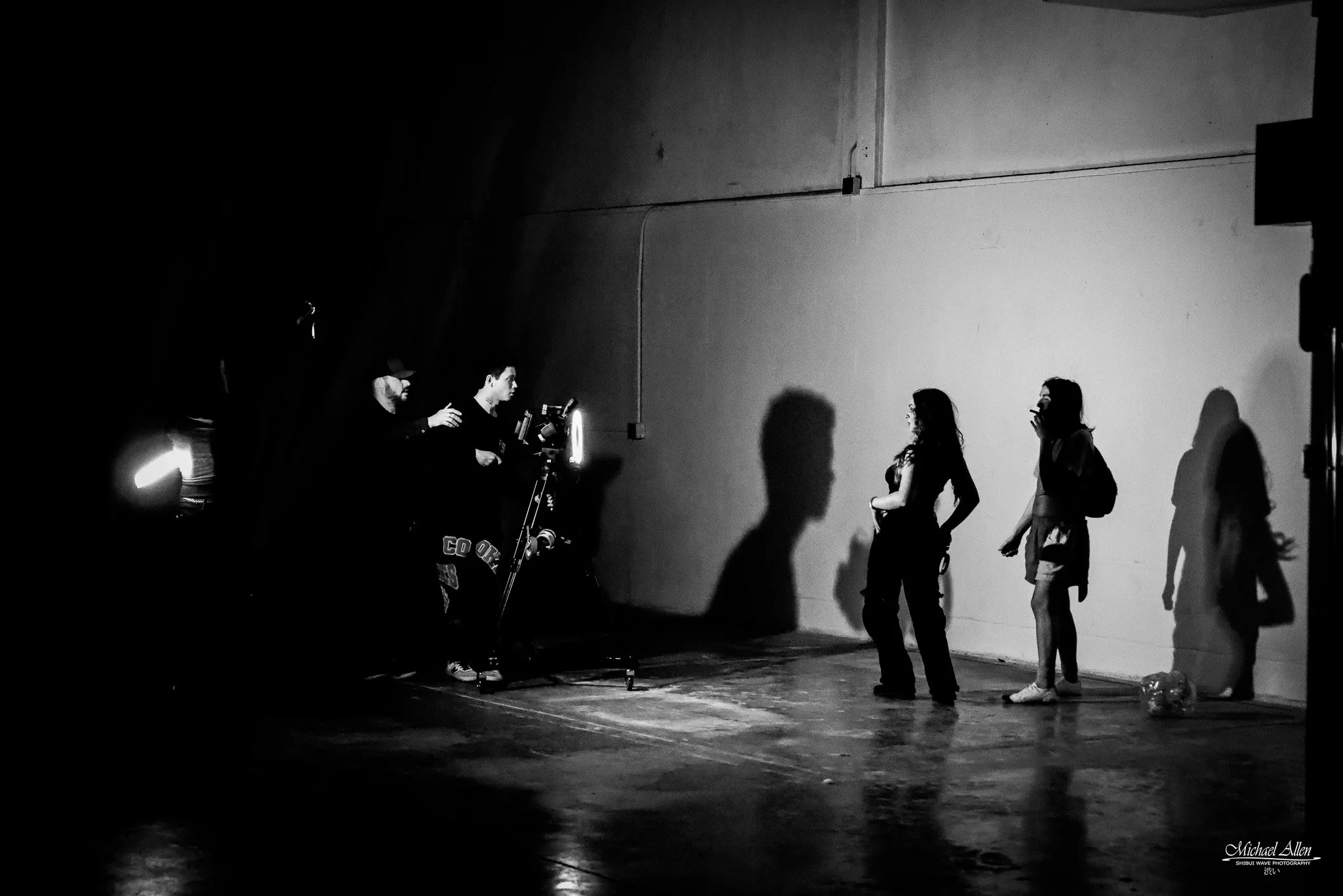 Black and white photo of a dance studio with three women in front of a plain wall and three men who are working with lighting equipment. The women are standing to the right, one in mid-step, while the men are on the left, adjusting a light. The scene is dimly lit, creating shadows on the wall.