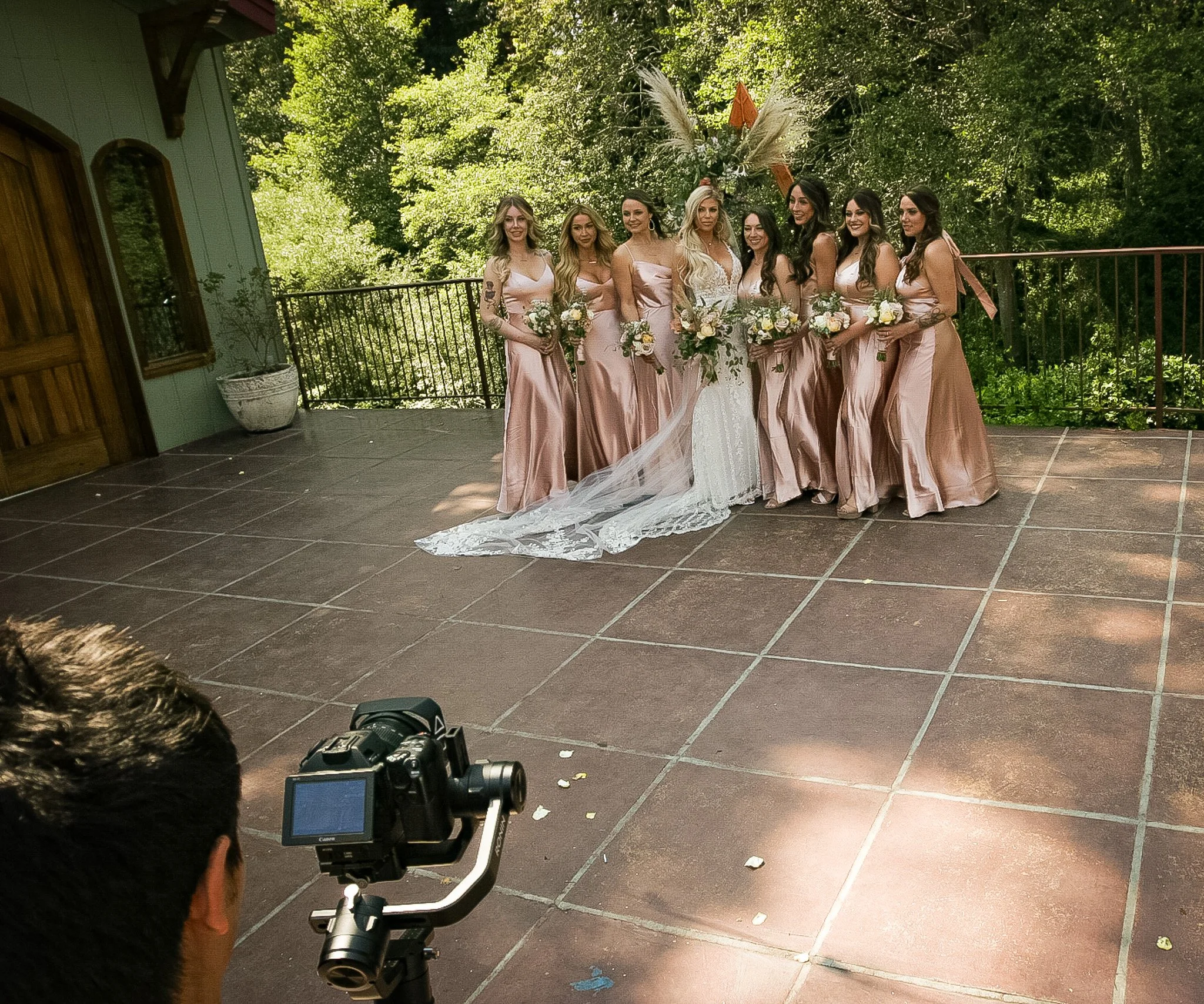 Group of women in peach-colored dresses holding bouquets, standing outdoors on a tiled patio with lush green trees background, during a wedding or special event.