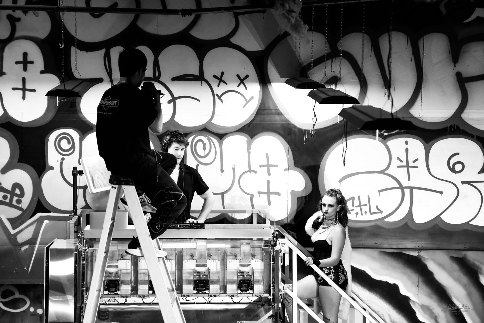 Black and white photo of three young people in a graffiti art studio. Two are on a platform with DJ equipment, one is standing on stairs, and a young woman with dark makeup and wavy hair is standing at the bottom, posing with her hand on the stair railing.