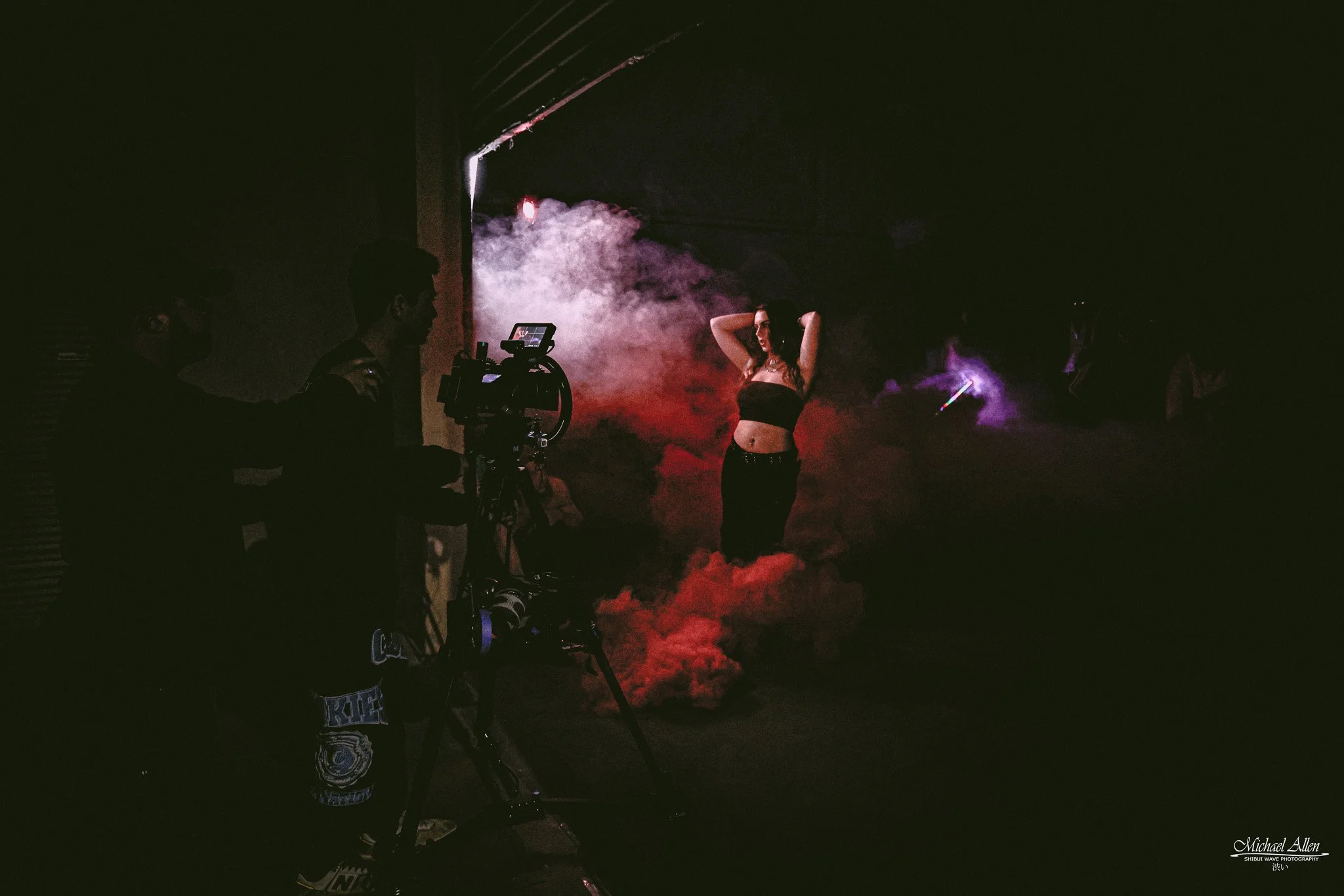 A woman standing amidst colorful smoke and fog, with photographers and camera equipment capturing her in a dark setting, possibly at a nightclub or photo shoot.