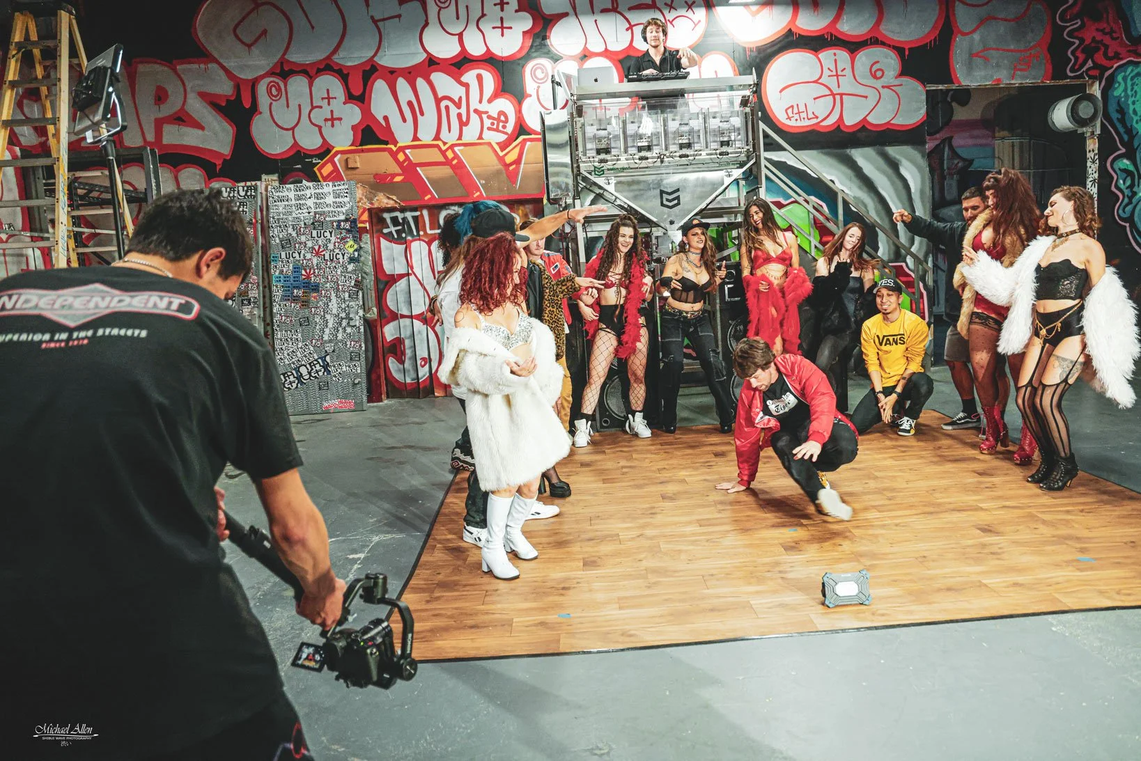 People in colorful costumes and outfits dancing and posing on a small wooden dance floor in an urban-style, graffiti-covered studio with a graffiti backdrop and a DJ at a turntable on elevated platform.