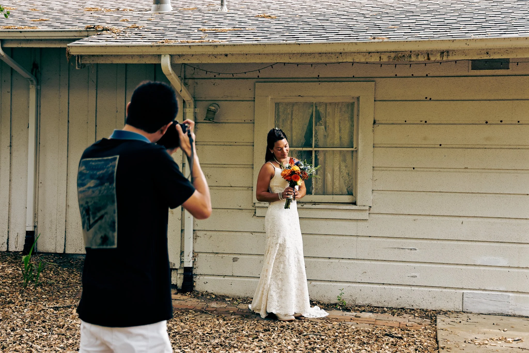 A woman in a white wedding dress holding a bouquet of flowers stands outside near a house with a wooden siding, as a man takes her picture.