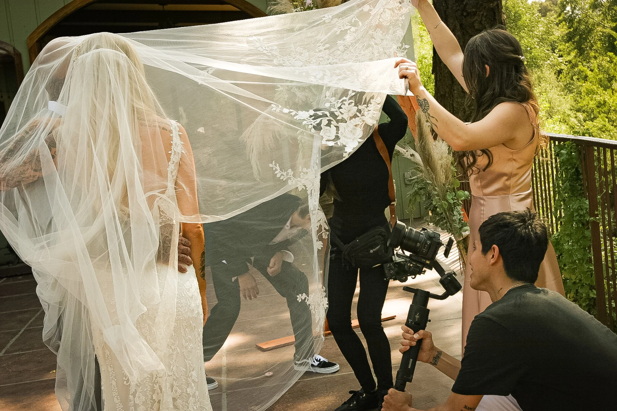 A bride with a veil standing outdoors during a wedding preparation, surrounded by her wedding attendants and photographers capturing the moment.