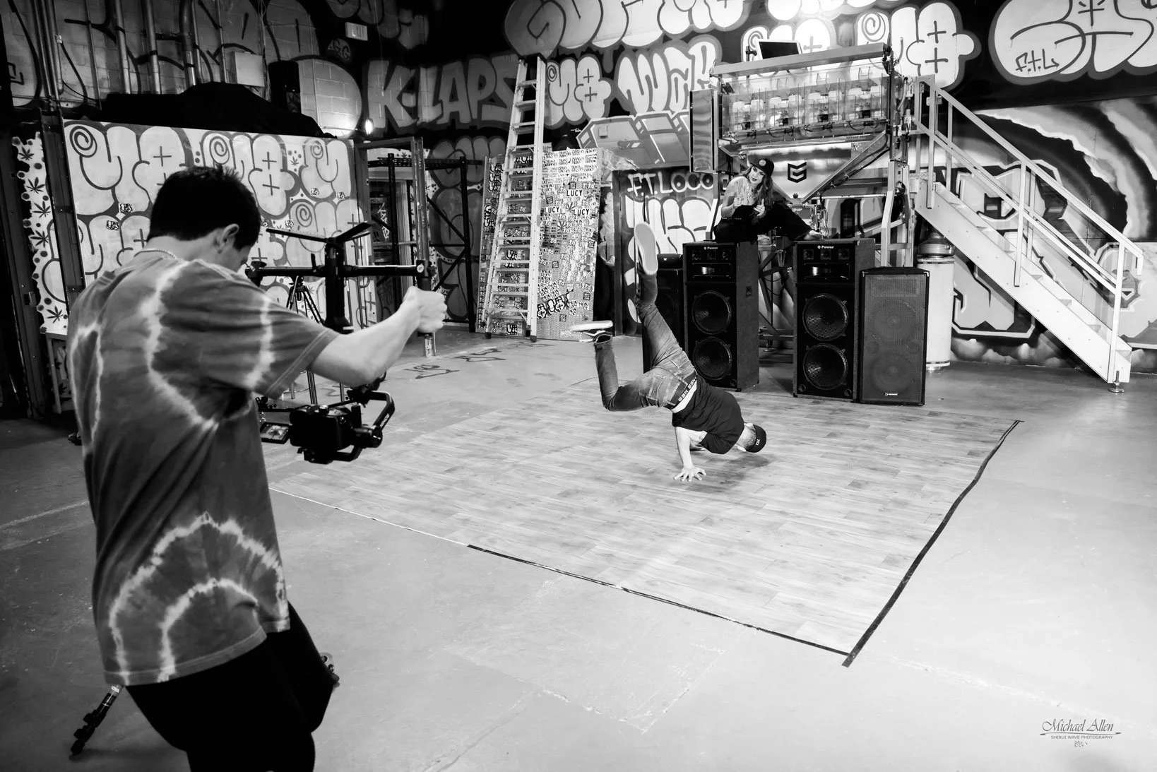 A person breakdancing on a wooden-floor dance studio, with another person recording using a camera mounted on a stabilizer, and a woman sitting on speakers in front of graffiti-covered walls.
