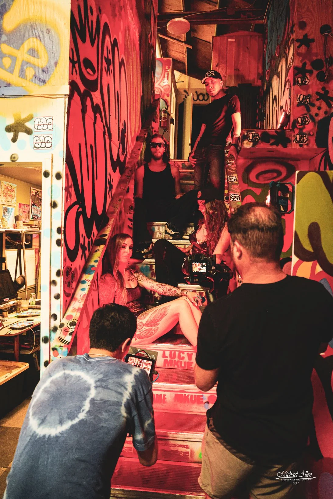 A group of people and artists on a colorful, graffiti-covered staircase. Some are seated and one standing, with the scene being photographed or filmed by two men in the foreground.