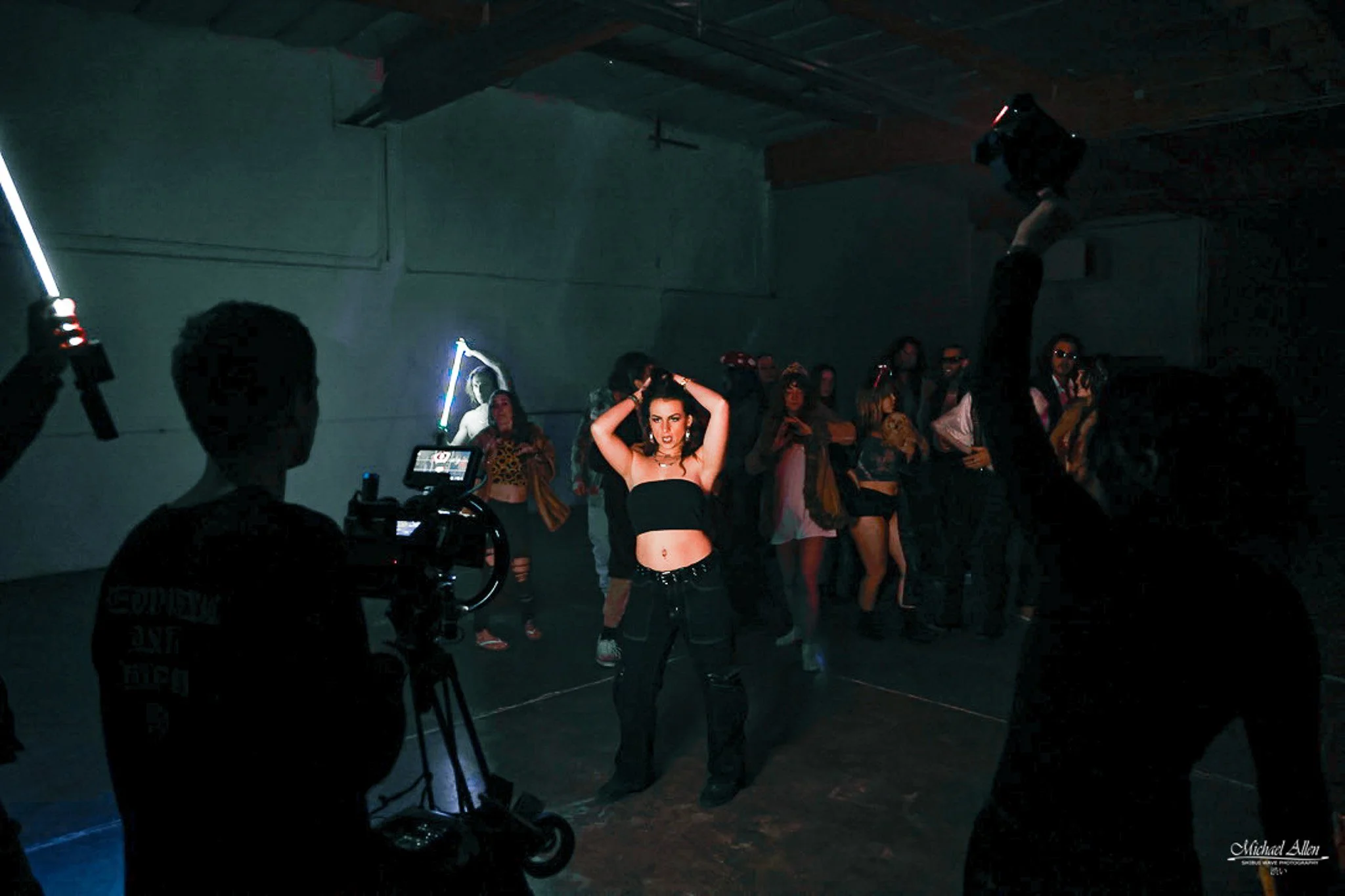 A woman in black clothes dancing in a dimly lit room, surrounded by a crowd, with a film crew filming her. She is raising her hands to her head, and the crowd is watching her performance.