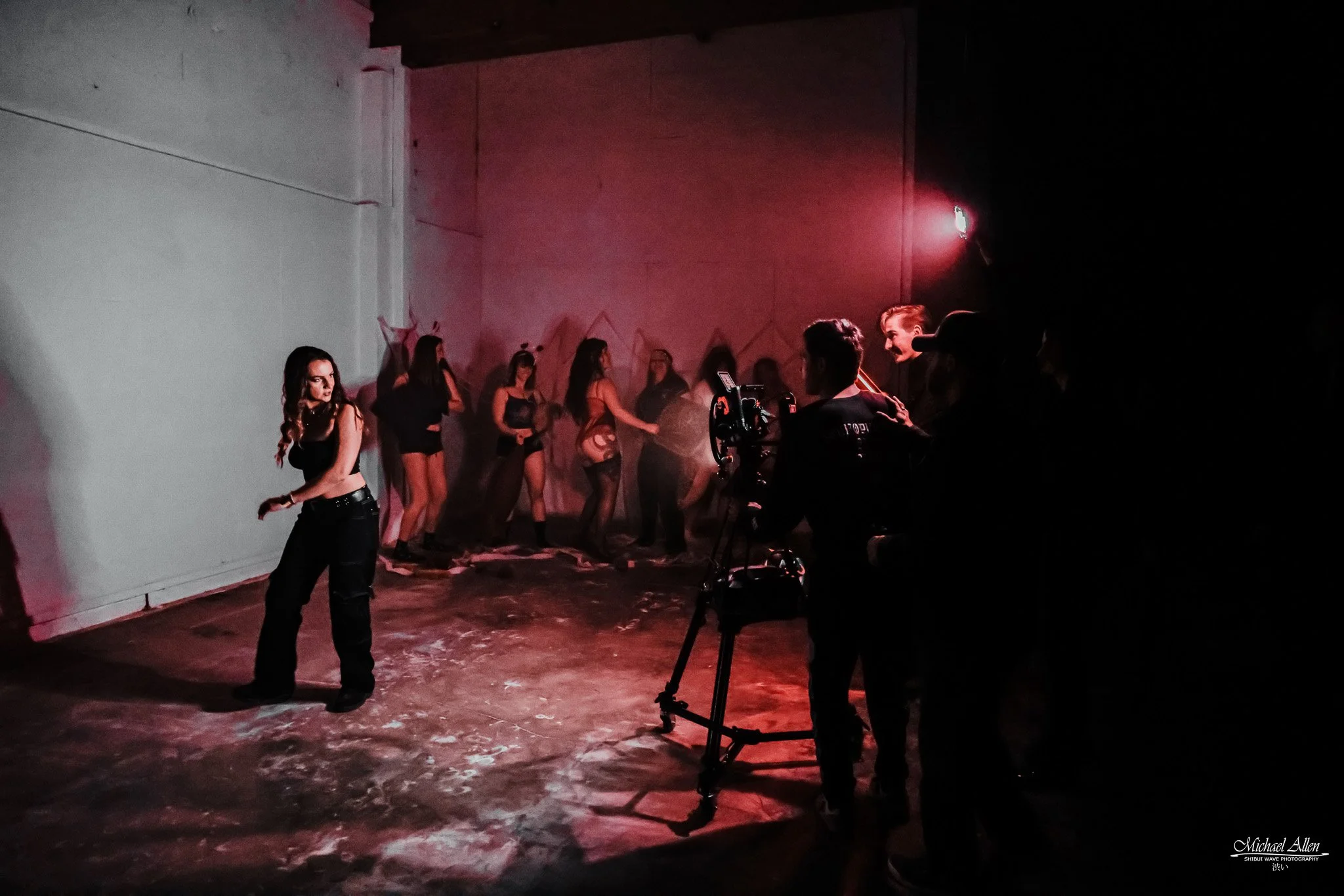 A group of women in black outfits and high heels are standing against a plain wall, while a crew films them with camera equipment in a dark, indoor studio with red and pink lighting.