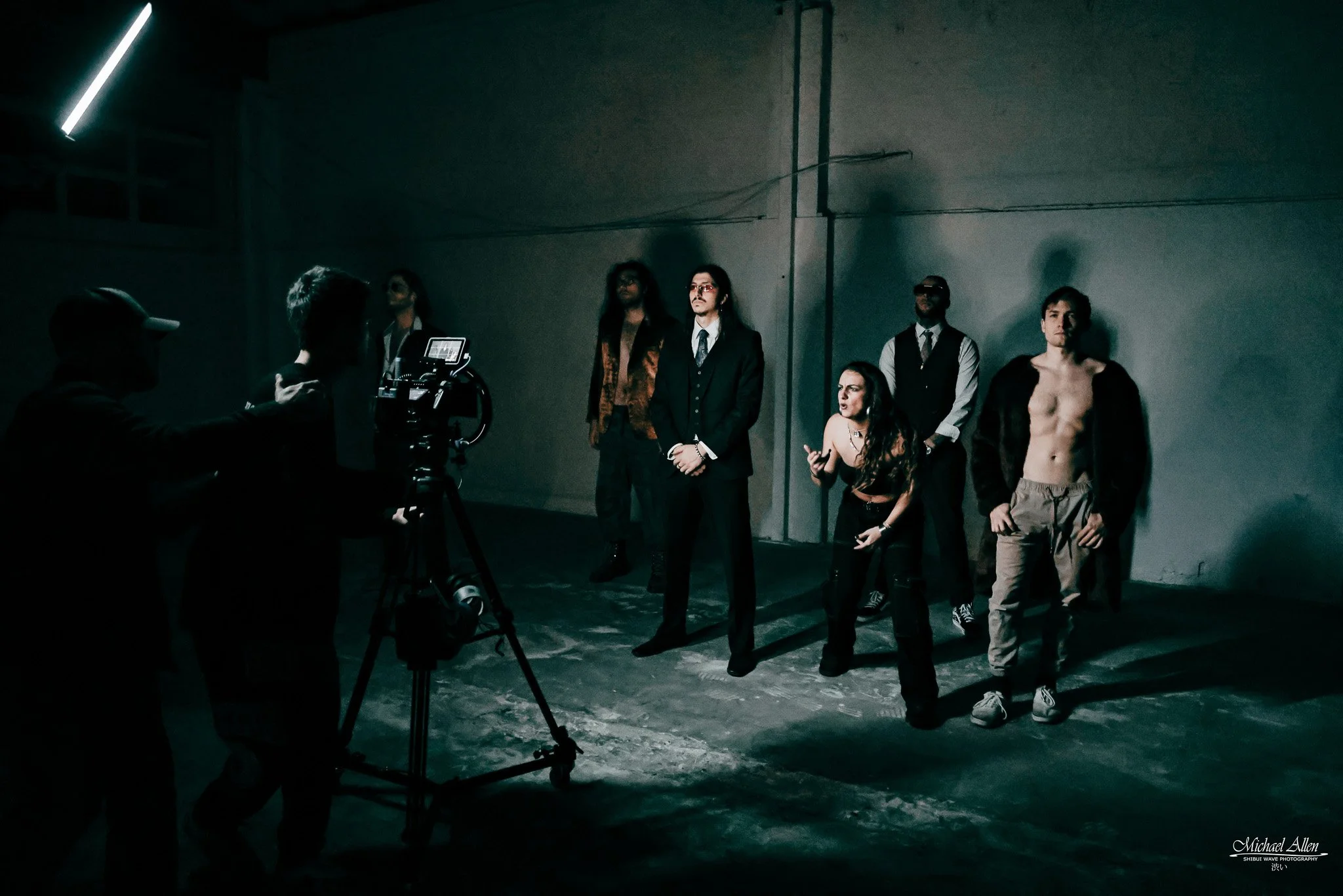 A group of six people standing in a dimly lit, industrial-looking space with concrete floors and a plain wall. One person is shirtless with an open jacket, another wearing sunglasses and a suit, and others in casual or semi-formal attire. A person operating a camera or filming equipment is in the foreground.