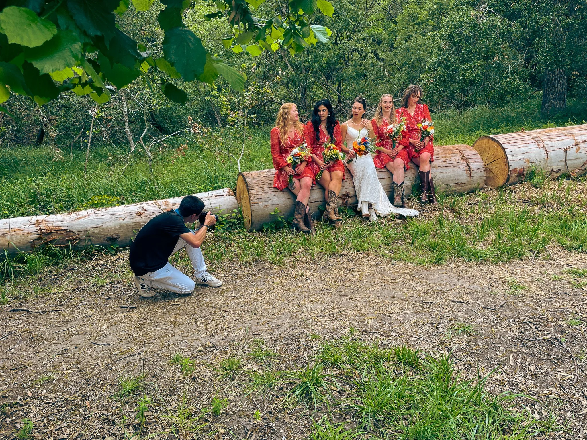 Group of five women sitting on a log in a forested area, holding bouquets of flowers, while a man kneels taking a photograph near them.