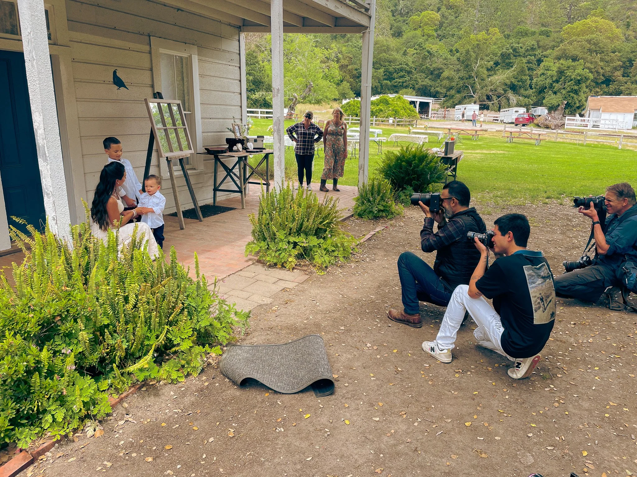 Two children and a woman at a sunny outdoor gathering, with photographers taking pictures of the children and woman, and a couple of people standing and talking nearby.