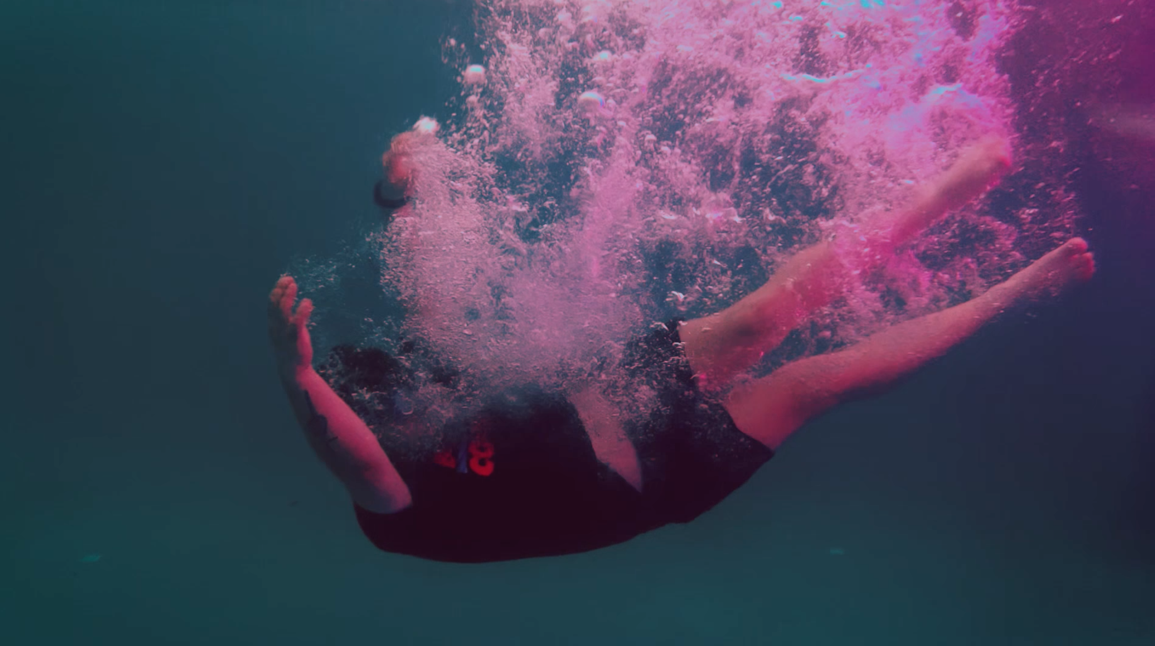 Person underwater with hands together, creating a splash of pink bubbles.
