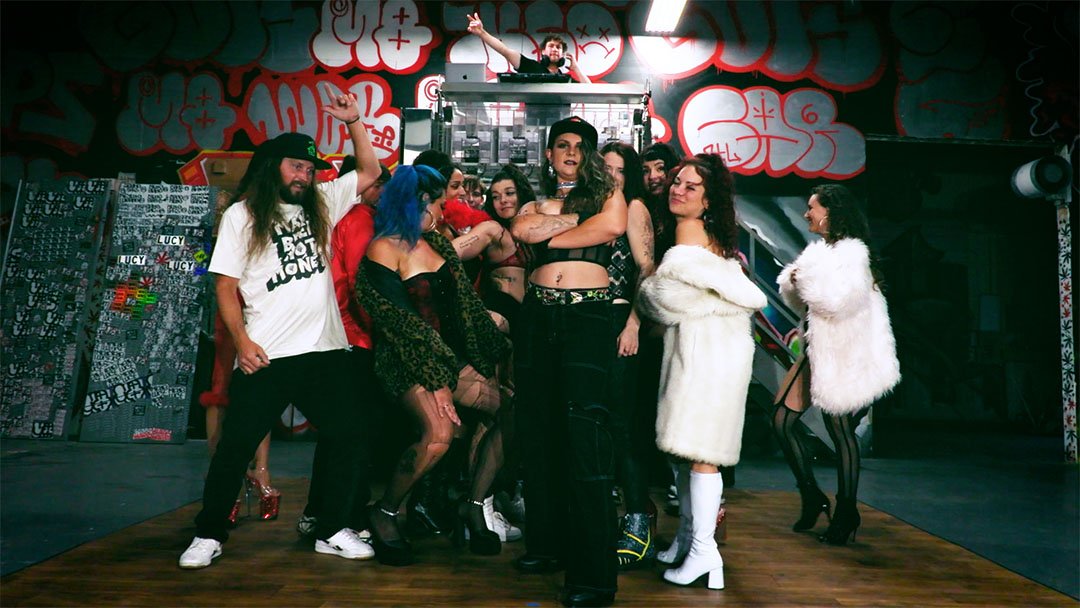 A group of people posing in a graffiti-covered indoor space. Some individuals are wearing colorful and fashionable clothing, including fur coats, fishnet stockings, and bold hairstyles. One person is DJing from a booth above the group.