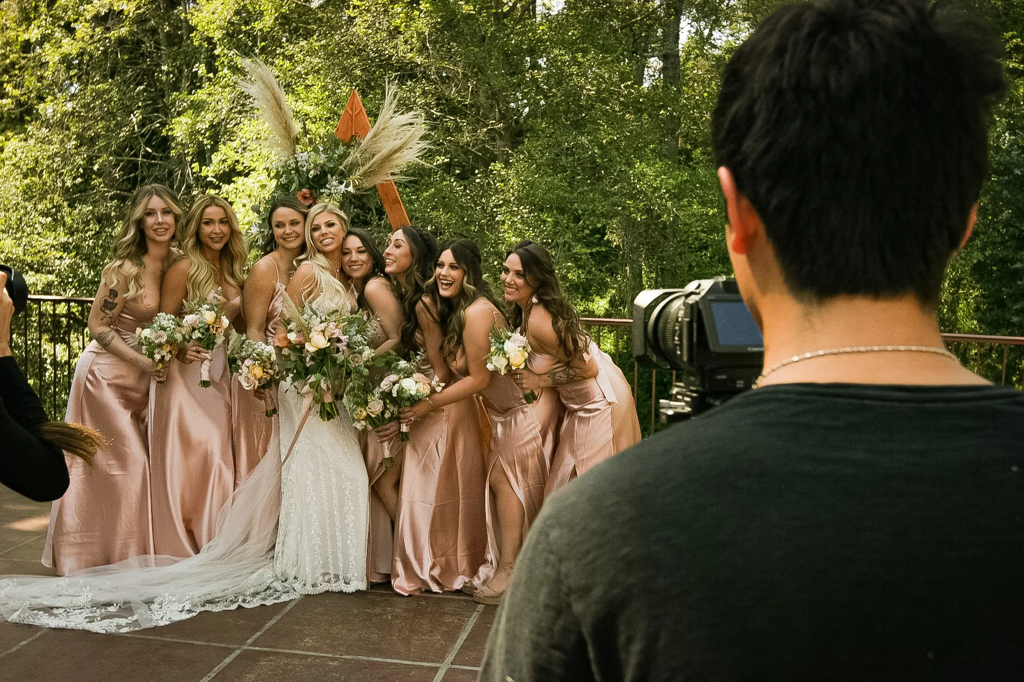 A group of women in matching pink dresses holding bouquets, posing for a photo in an outdoor setting with trees, while a person with short dark hair operating a camera is in the foreground.