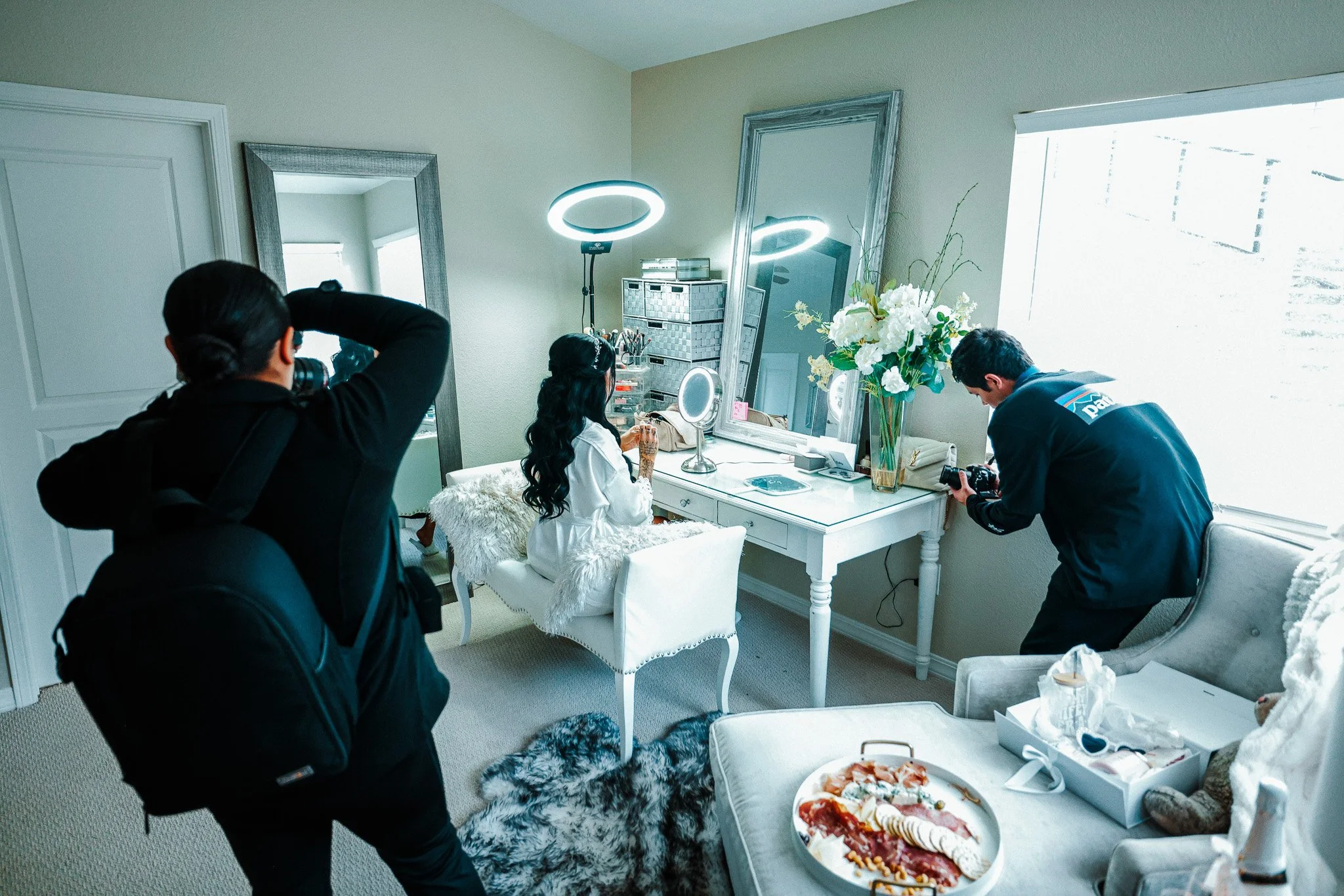 People preparing for a photoshoot in a well-lit bedroom. One person is sitting at a vanity, while others set up lighting and cameras.