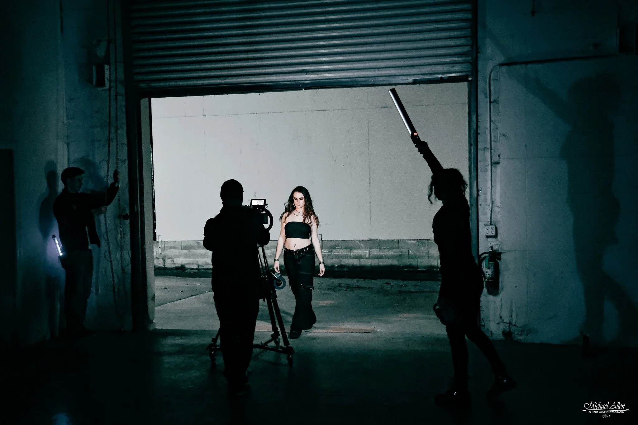 A woman in black clothing standing indoors, being filmed by a camera crew in a dimly lit industrial setting with a large, partially open metal roll-up door behind her.