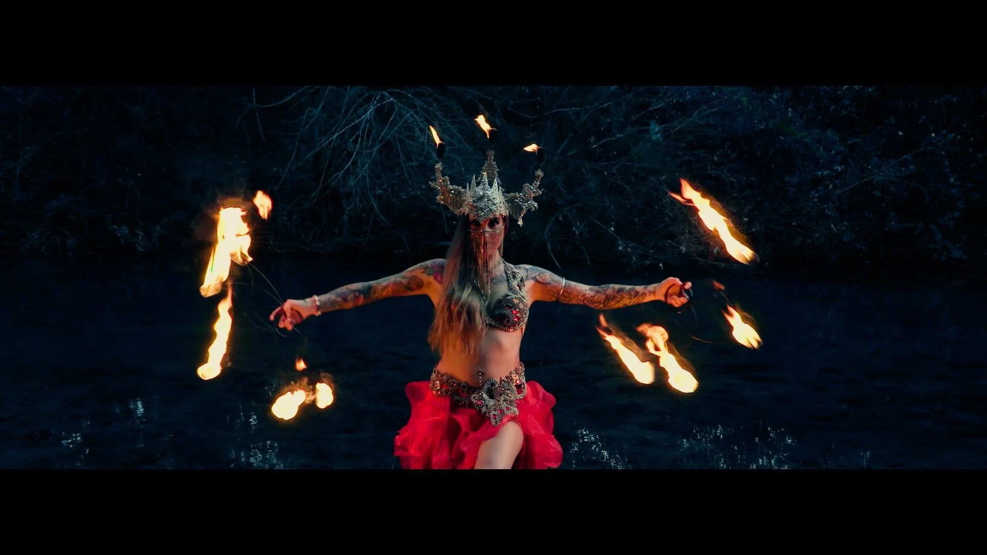 A woman dressed in a red skirt, ornate jewelry, and a crown performing fire dancing by a dark body of water at night.