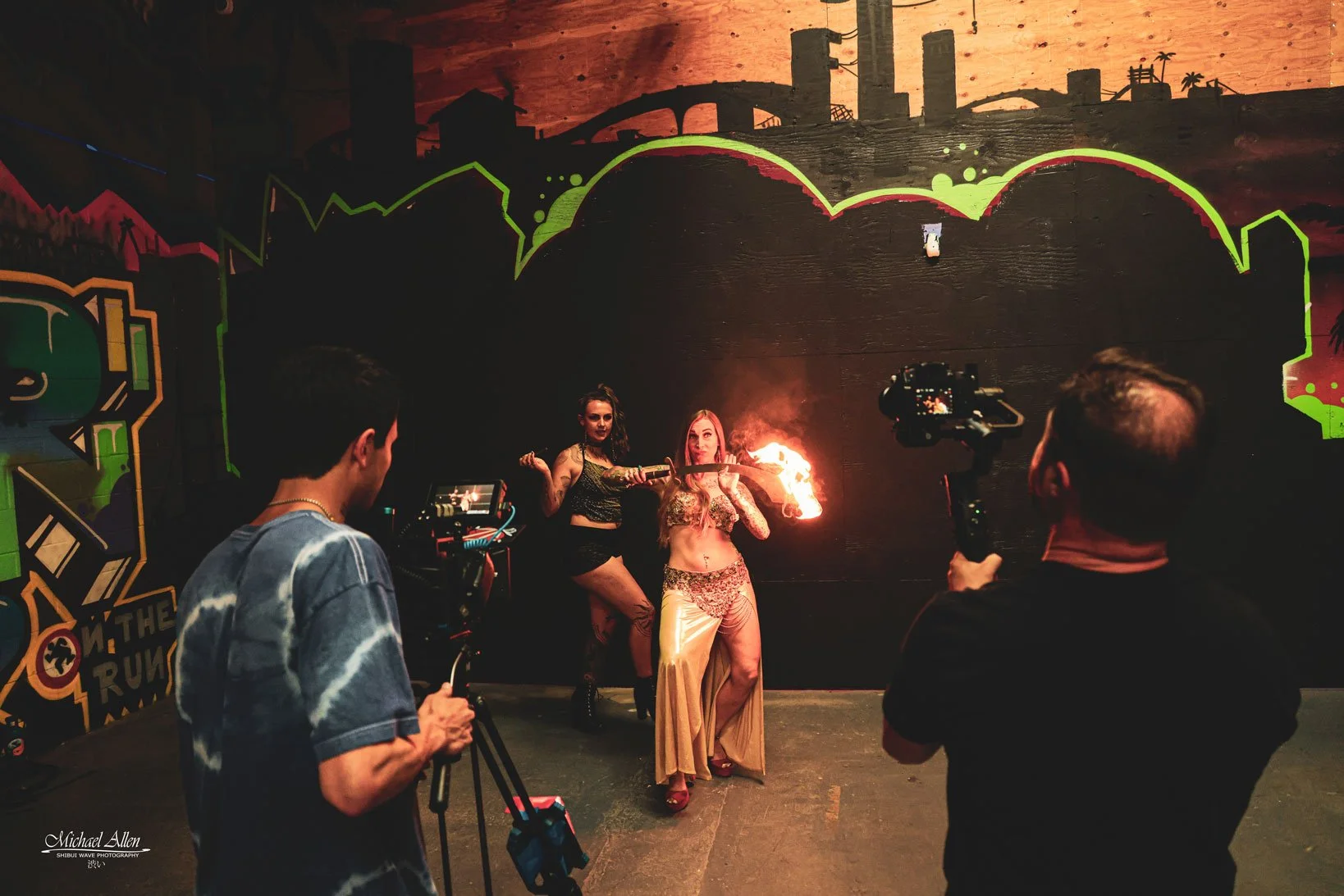 Two women performing a fire dance in front of a dark wall with cityscape murals, with one woman holding a flaming torch and the other holding a sword. Two men are filming the performance, one with a camera on a tripod and the other holding a small camera, in a dimly lit indoor venue with graffiti-style artwork on the walls.