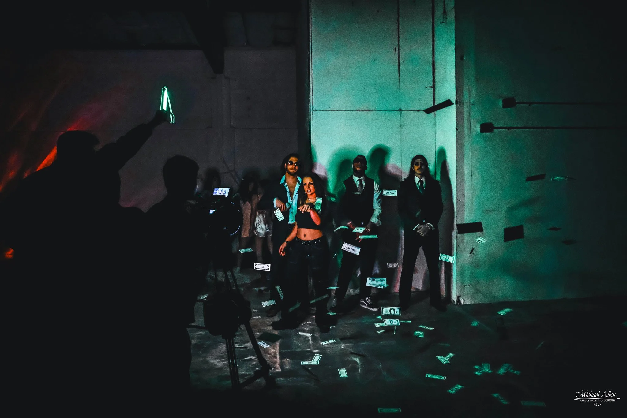 People posing with dollar bills falling around them in a dimly lit room with industrial walls and green lighting.
