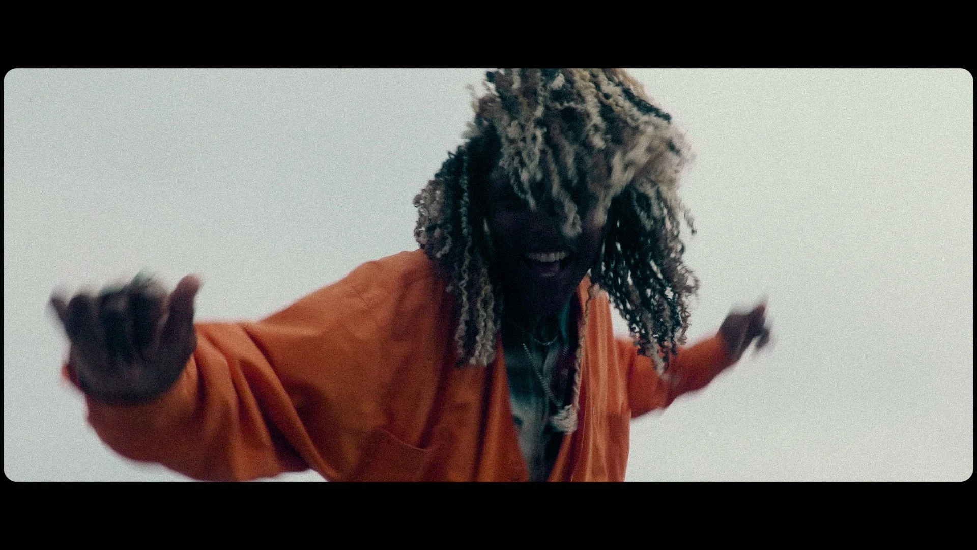 Person with blonde and black dreadlocks smiling and reaching out toward the camera, wearing an orange jacket.