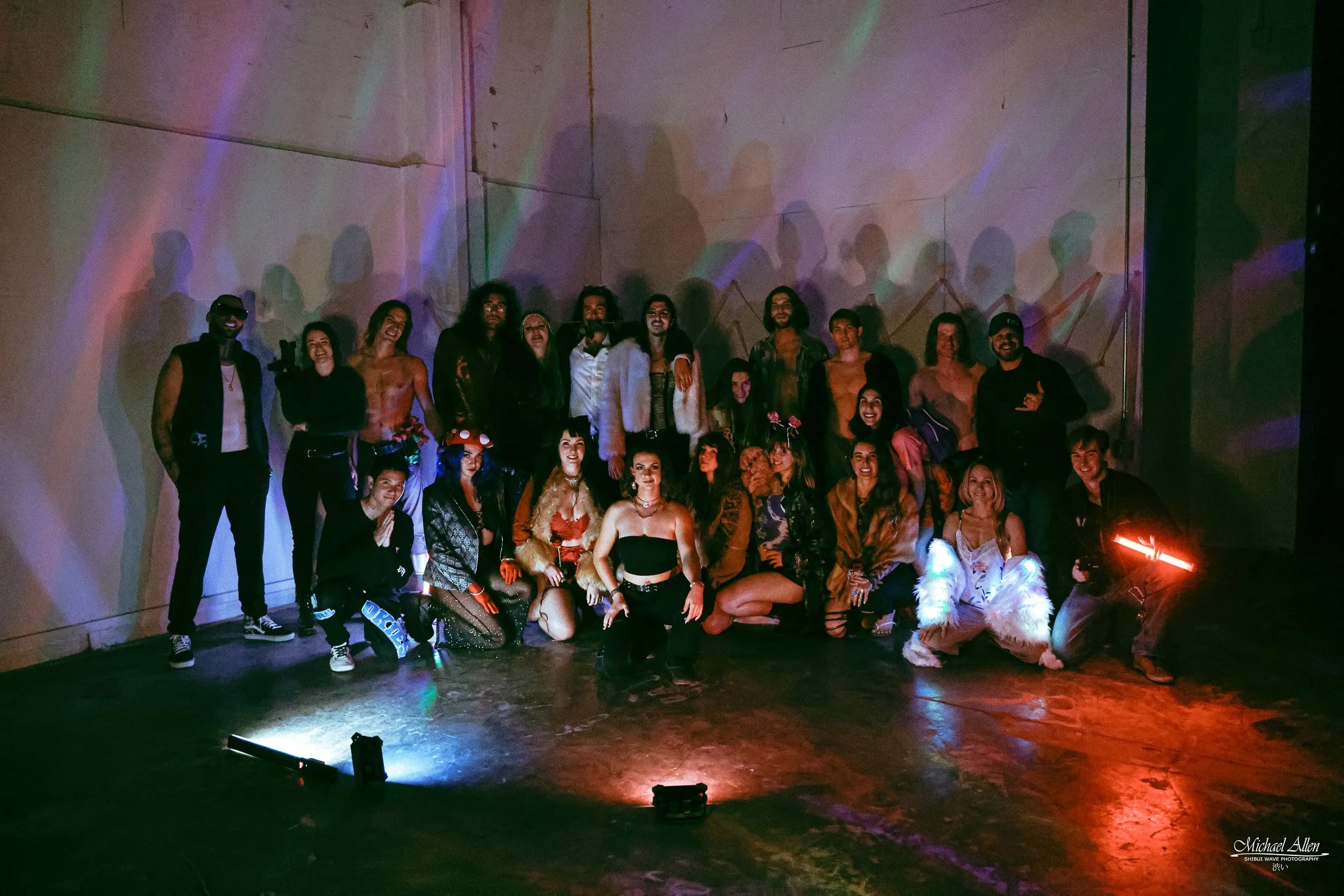 Group of people posing together in a dimly lit room with colorful lights projecting shadows on the wall behind them.