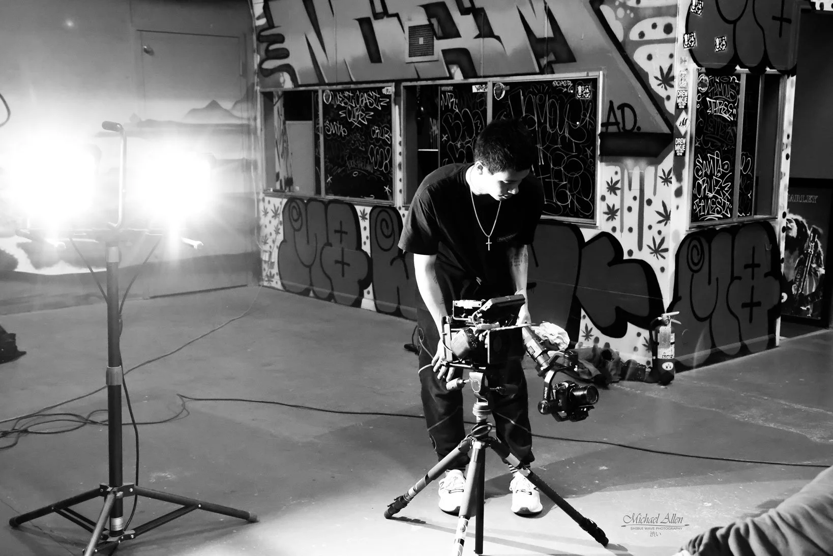 A person operating a camera on a tripod in a studio with graffiti art on the walls. Bright light fixture on stands is visible to the left.
