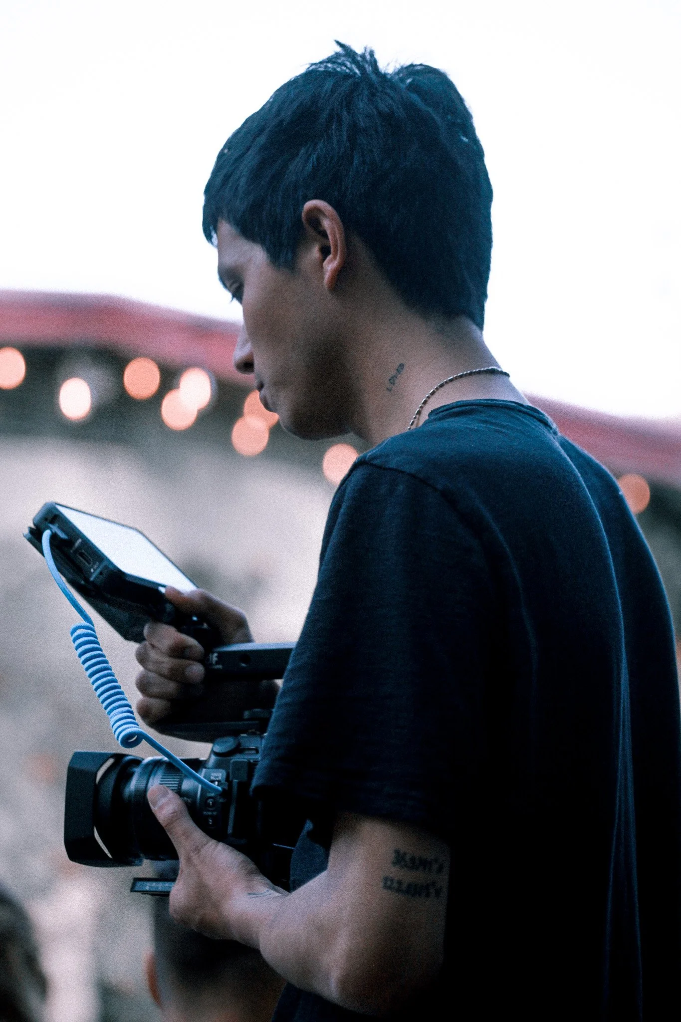 Person with short dark hair holding a camera with a strap, wearing a black shirt, and tattoos on their arm, in an outdoor setting at dusk.