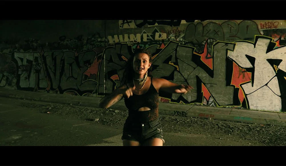 A woman dressed in black, wearing necklaces, smiles while running or dancing in front of a graffiti-covered wall in a dark, urban setting.
