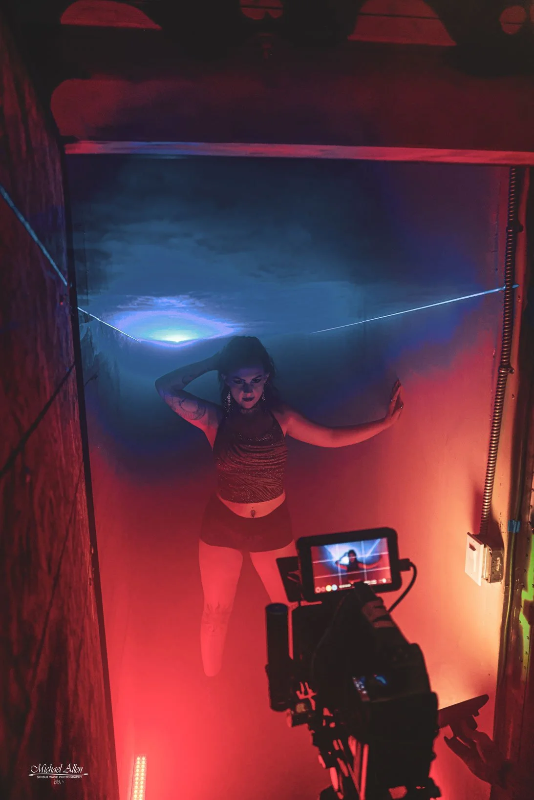 A woman with tattoos and dark makeup poses in a small, colorful room with red and blue lighting, while a camera records her from a lower angle.