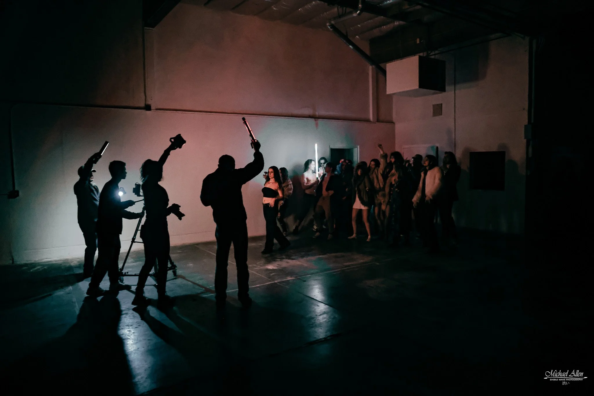 Dark indoor scene with people gathered, some holding phones or lights, and a few holding swords or sticks, with a large blank wall and ceiling beams in the background.