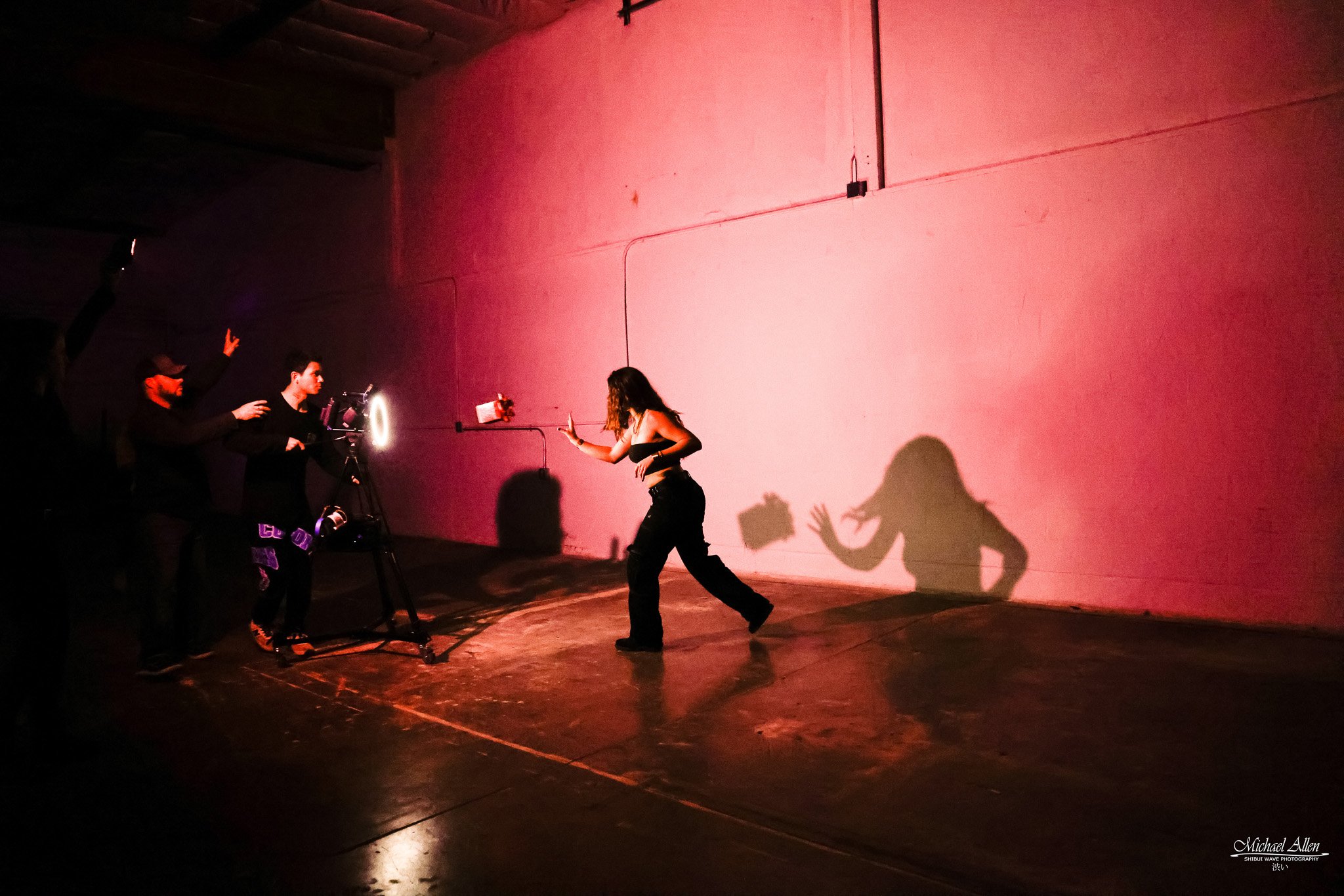 A woman dancing in front of a pink wall with shadows, while behind her, a crew films her with a camera on a tripod. The scene is dark with colorful lighting.