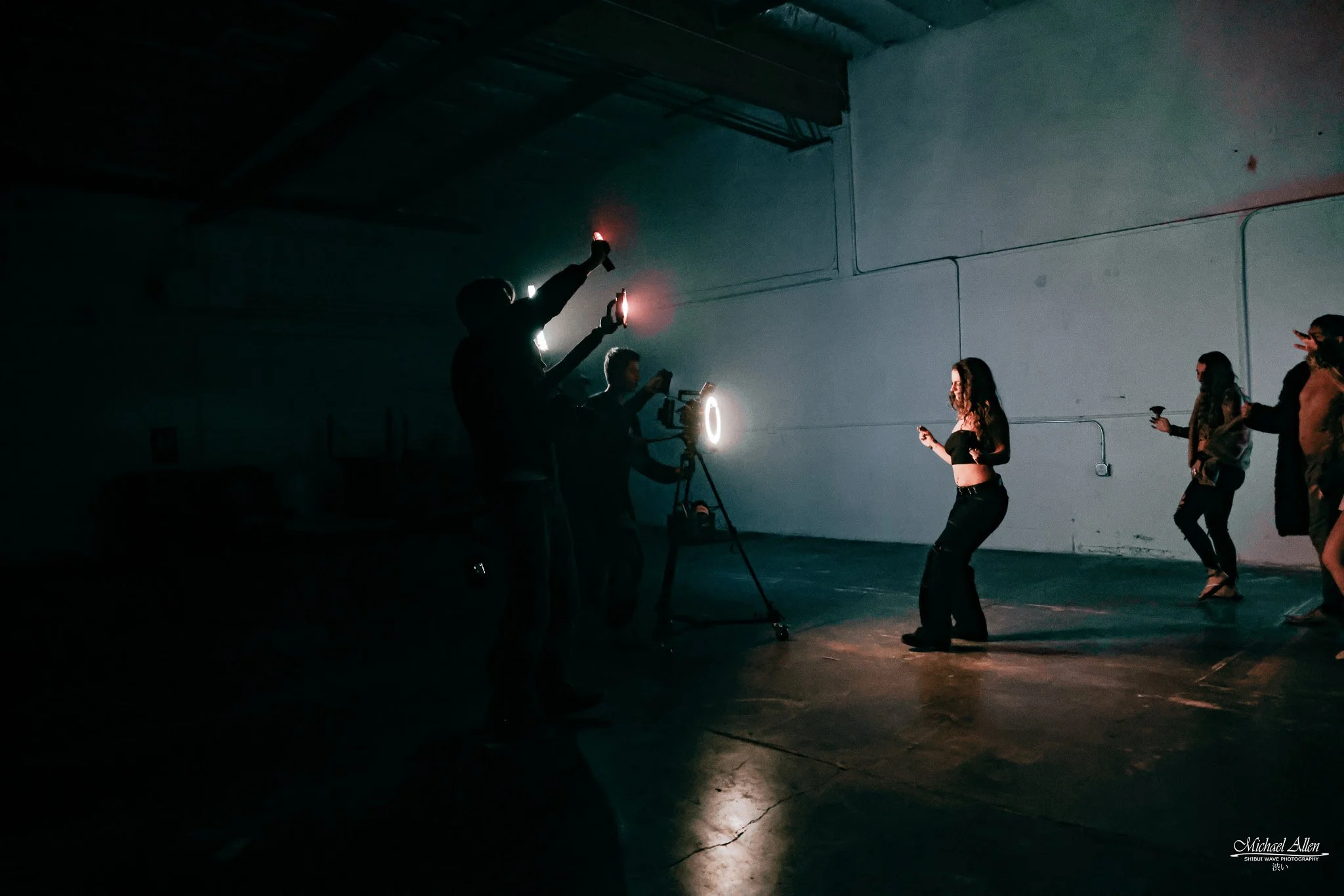 A dimly lit indoor setting with people, some holding phones and lighting equipment, and two women dancing in the background, illuminated by professional lighting equipment.