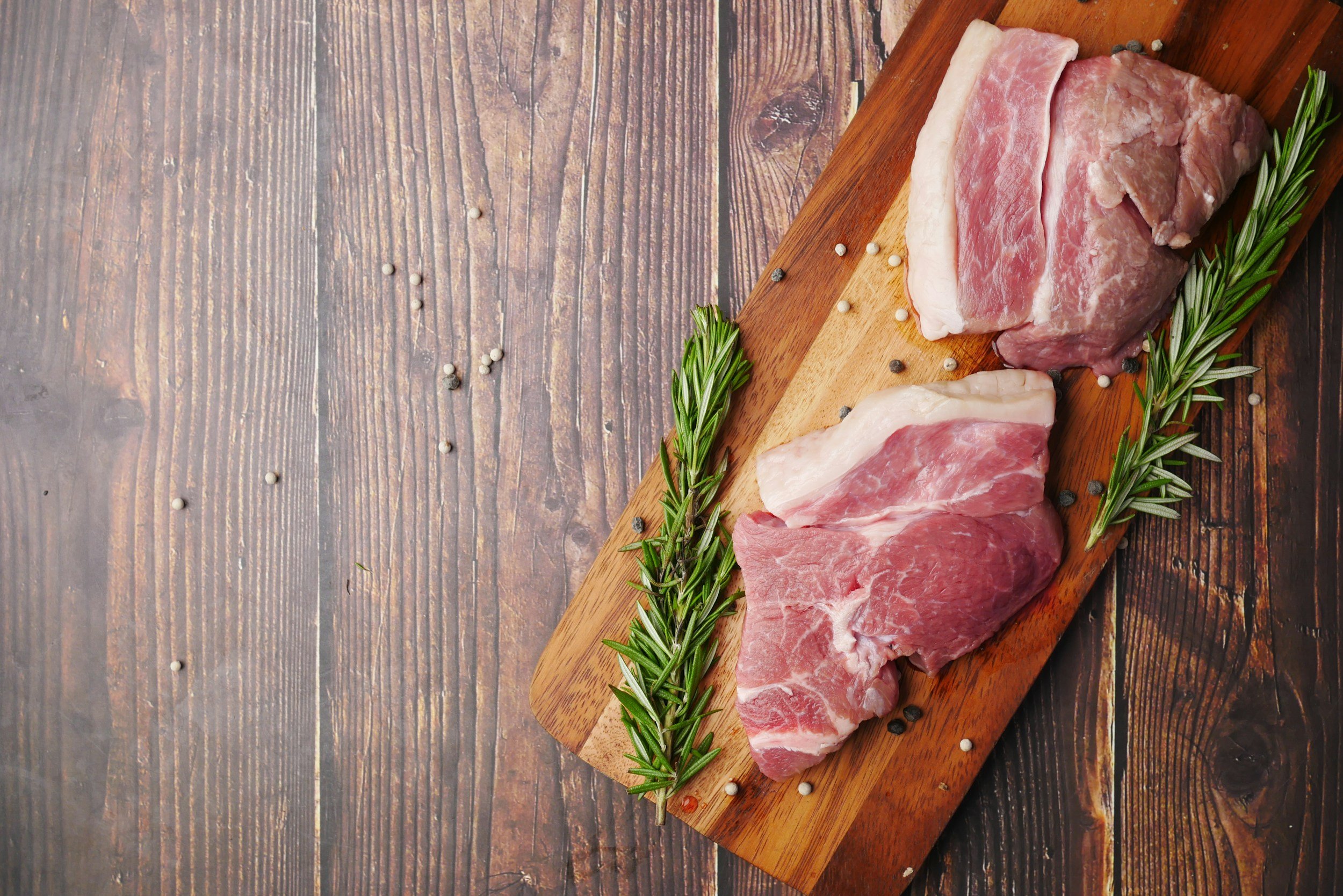 Raw pork chops with rosemary and peppercorns on a wooden cutting board.