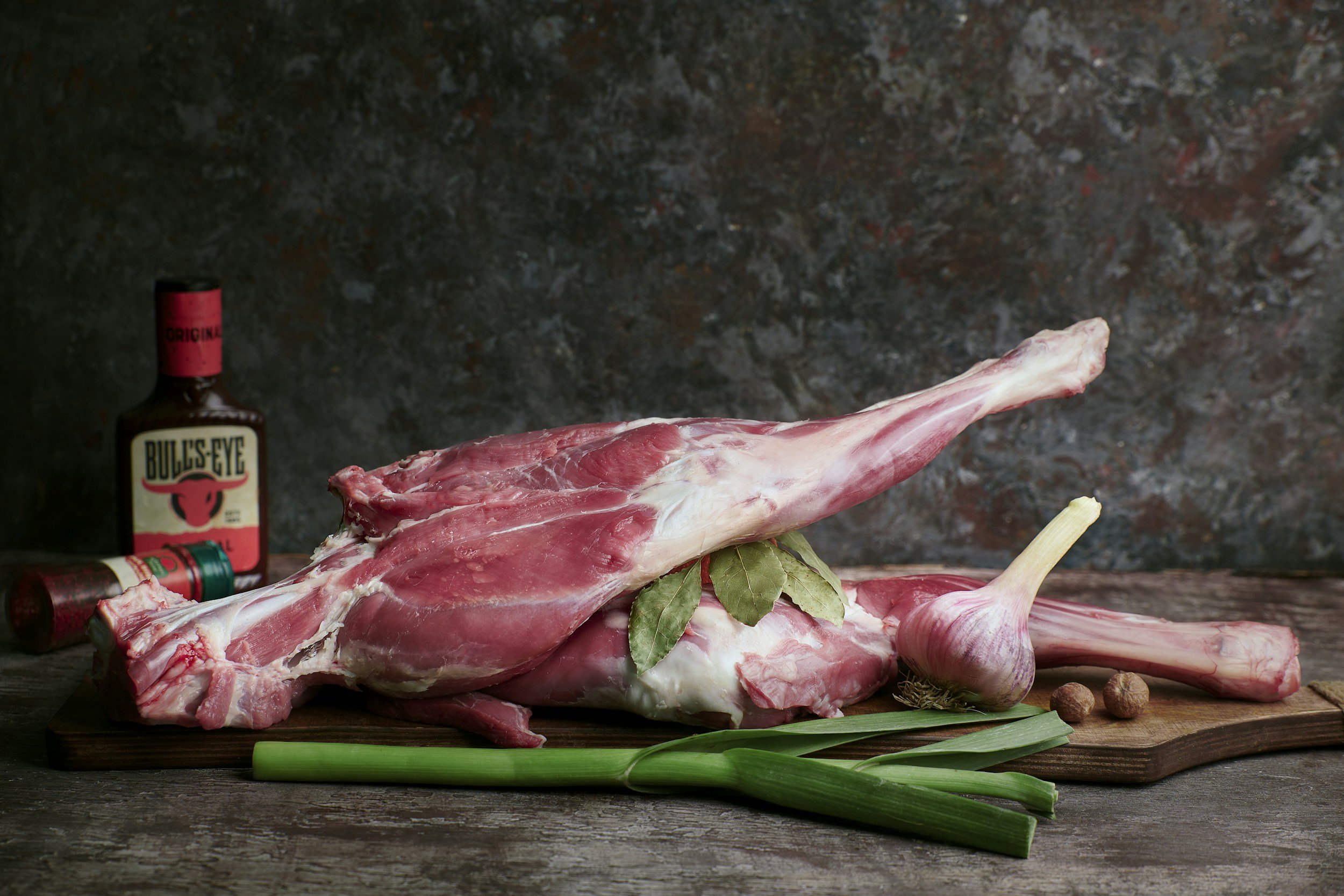 Raw lamb leg on cutting board with garlic, green onions, bay leaves, spices, and sauce bottle.