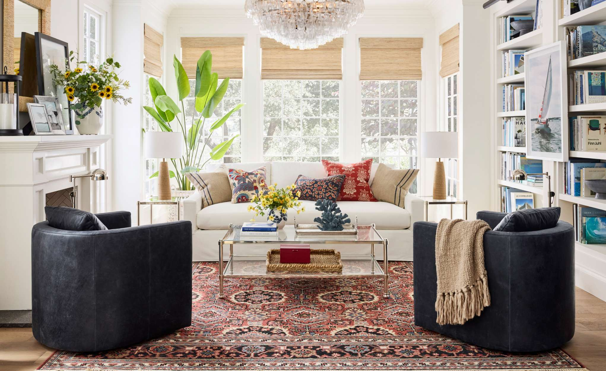 modern coastal living room with white sofa, blue leather chairs, traditional fireplace and traditional red and blue rug. This room has built in bookshelves and bamboo shades on the windows.