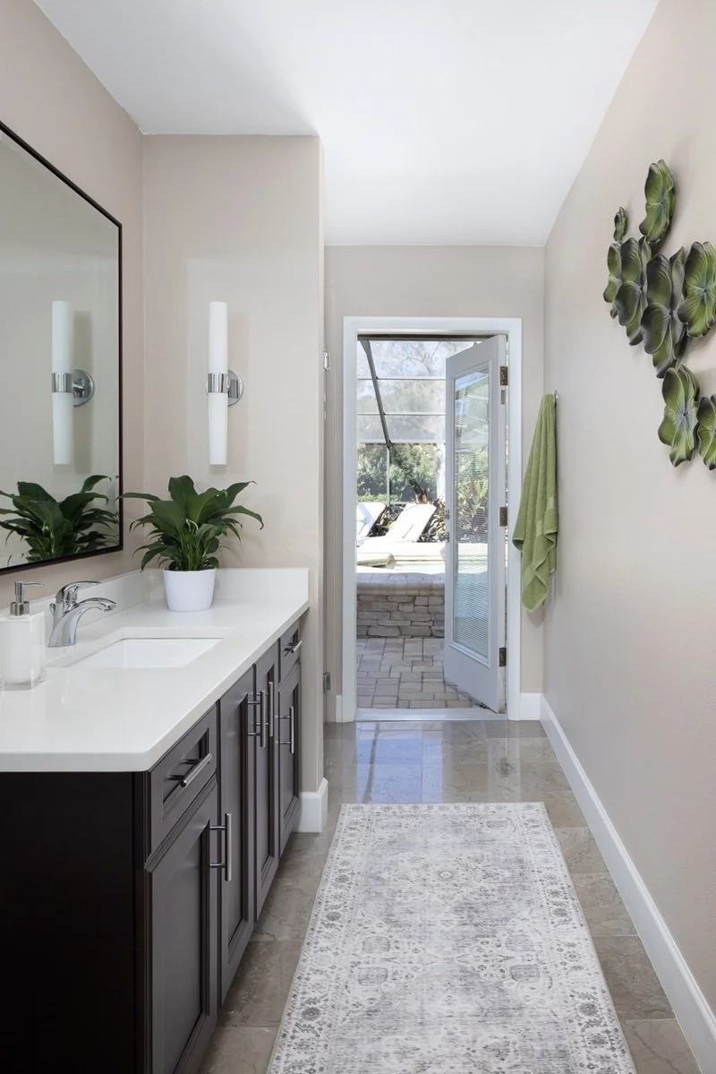 Warm white and taupe pool bath with open door leading out to stone pavered lanai, decorated with chrome fixtures and green accents in Palm Harbor, FL.