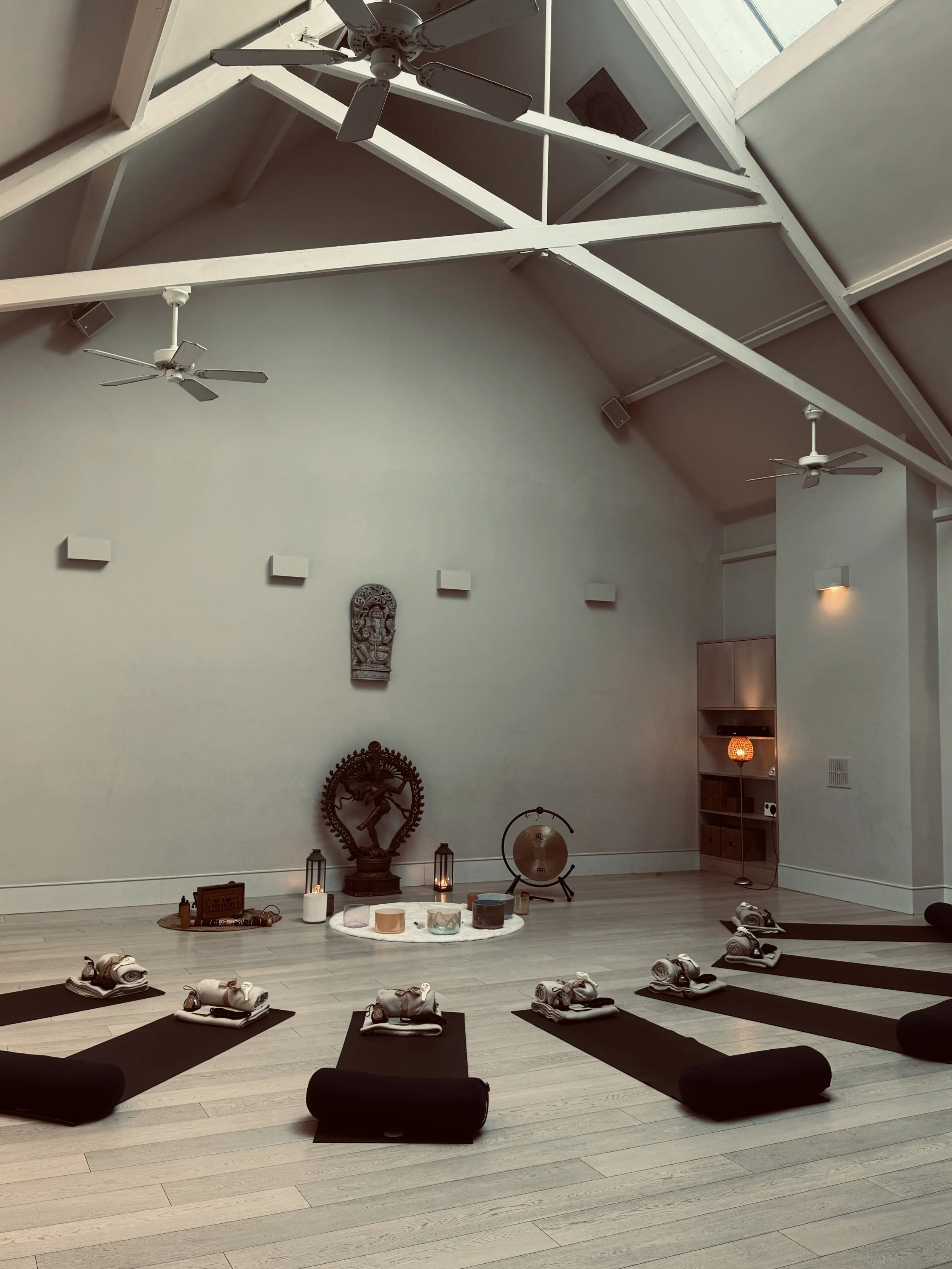 Indoor yoga or meditation space with black mats, rolled towels, candles, and decorative items arranged on the floor. The high ceiling features exposed white beams, ceiling fans, and skylights allowing natural light. The wall has a small sculpture and minimalist wall art. A soft ambient light illuminates part of the room.