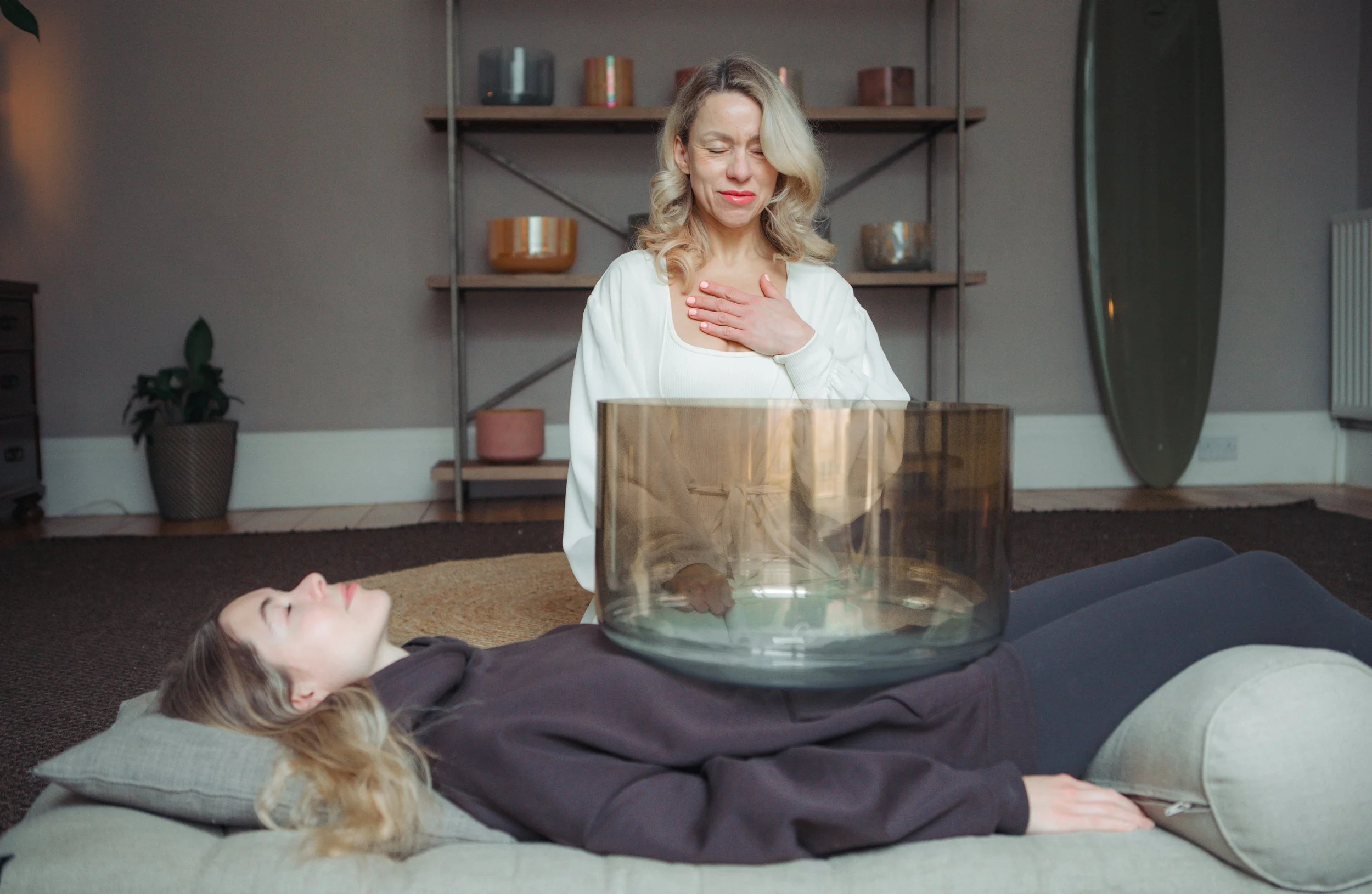 A woman is lying on a mat with a large singing bowl on her stomach, while another woman sits nearby with her hand on her chest, appearing to be participating in a spiritual or healing session.