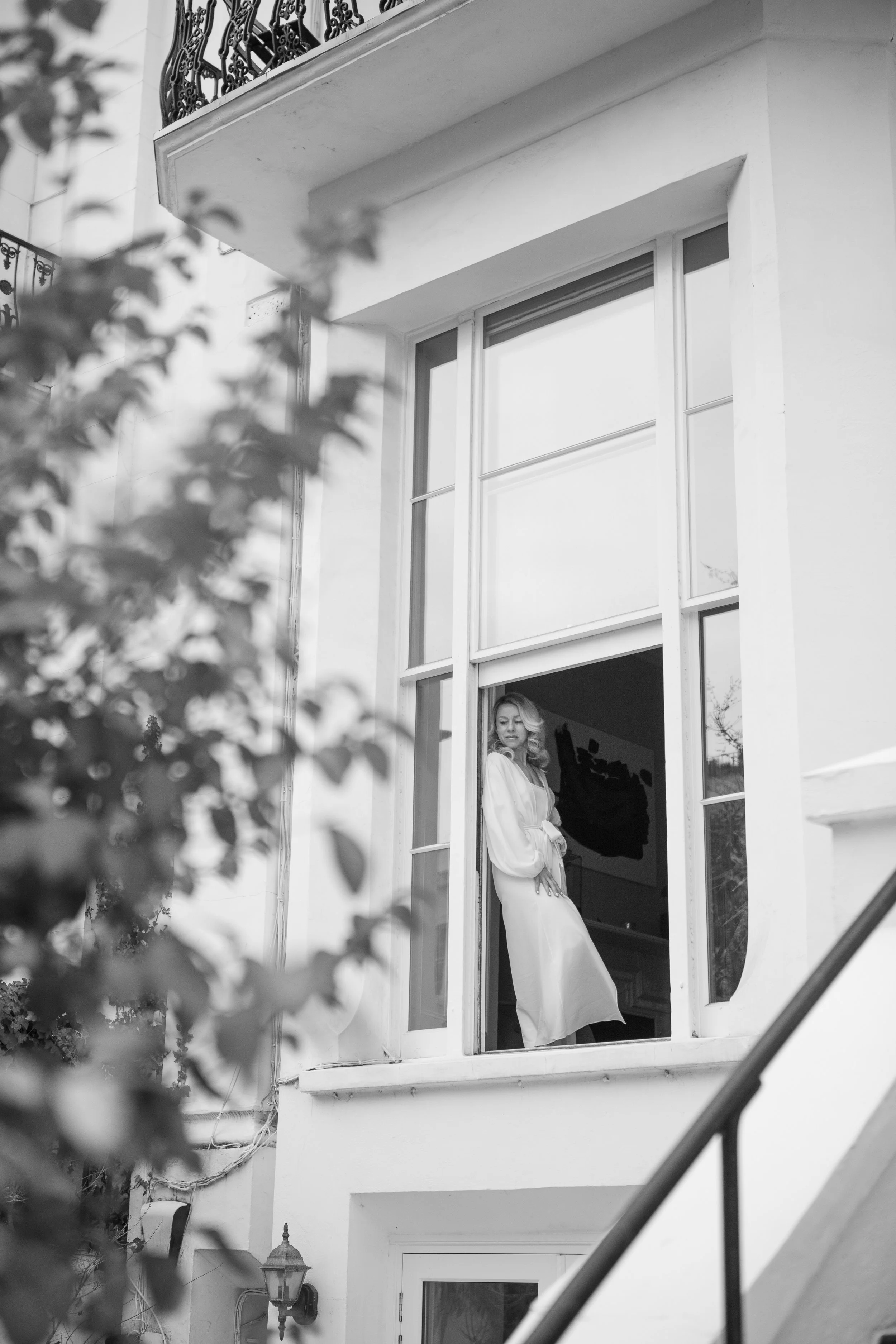 A woman with blonde hair and white clothing standing by an open window of an apartment building, looking out.