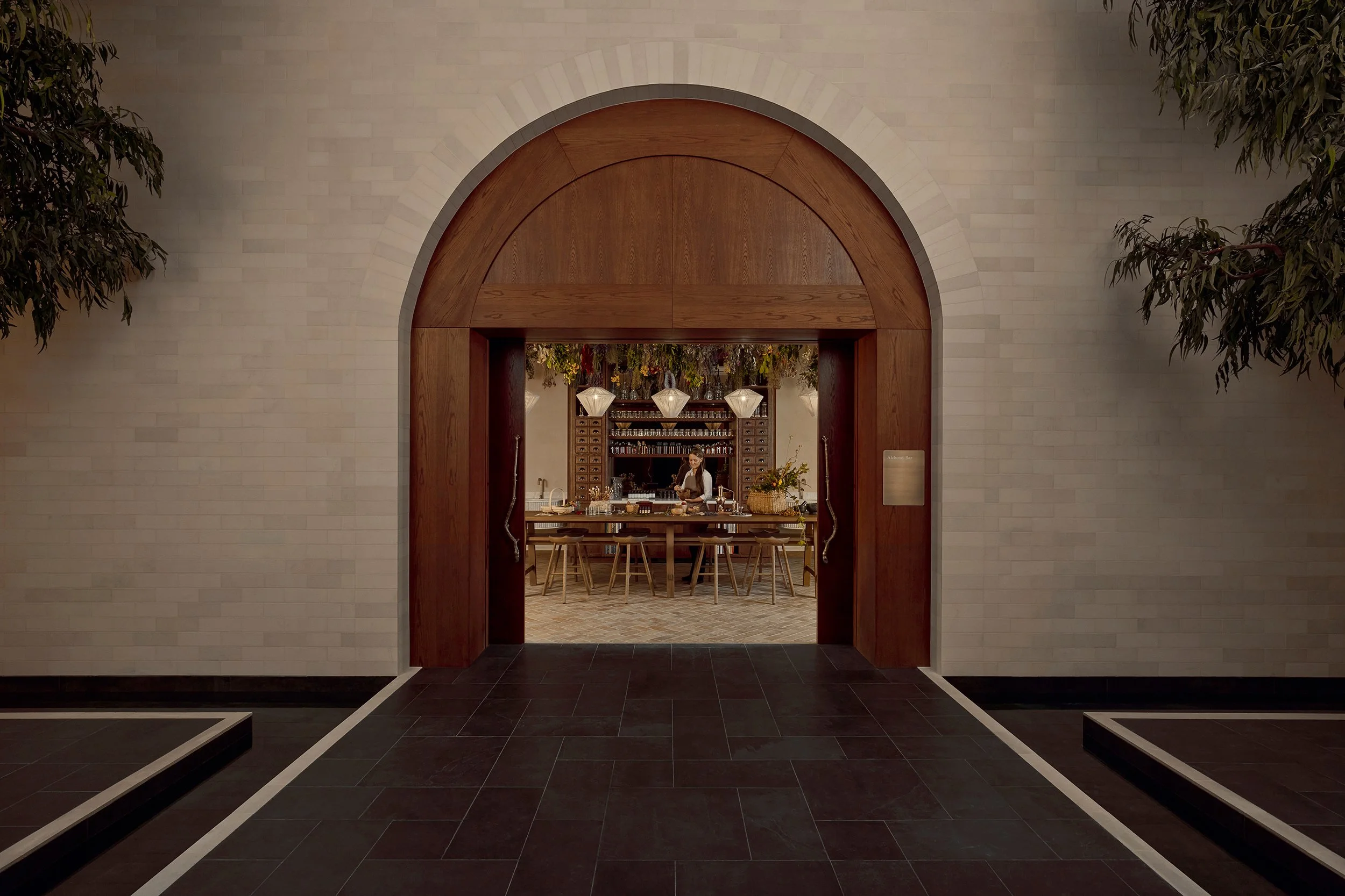 Interior view of a restaurant or cafe through a large arched doorway, with a bartender behind the bar, and pendant lights hanging from the ceiling, marble floor, and lush greenery on sides.
