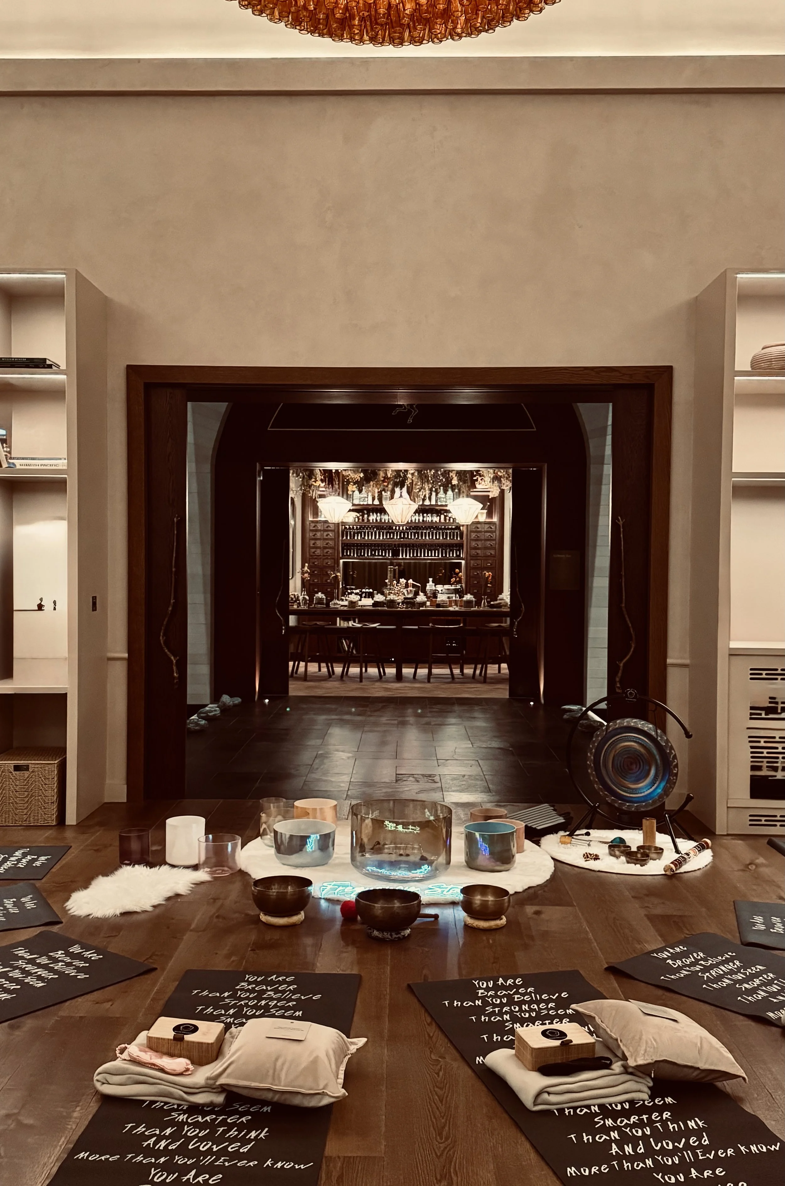 Elegant dining room with a long wooden table set with wine glasses, plates, and candles. A vase of flowers sits in the center. The background features a dark wood bar with shelves of liquor bottles and glass jars, and decorative hanging plants and lamps overhead.