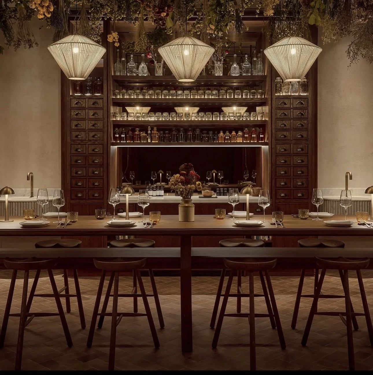 Elegant dining room with a long wooden table set with wine glasses, plates, and candles. A vase of flowers sits in the center. The background features a dark wood bar with shelves of liquor bottles and glass jars, and decorative hanging plants and lamps overhead.