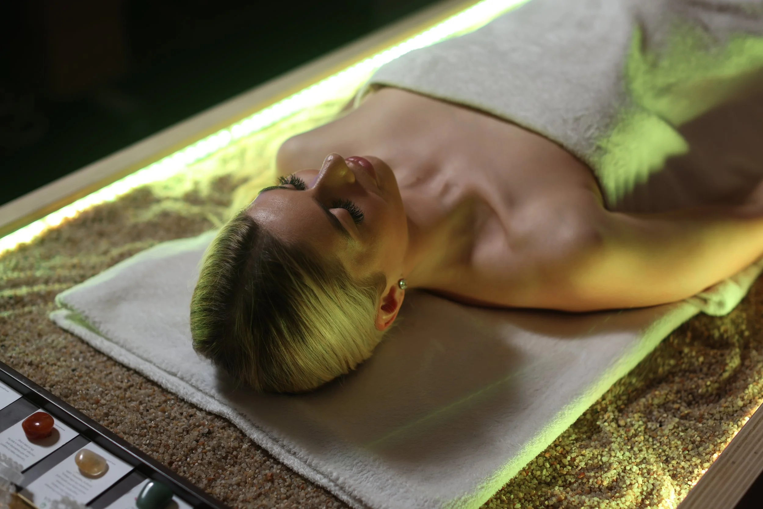 Person lying on a spa bed covered with a towel, illuminated with green light, with sand underneath.