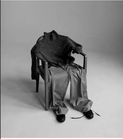 A surreal image of empty clothes arranged on a chair forming the shape of a sitting person with a sweater, pants, and shoes.