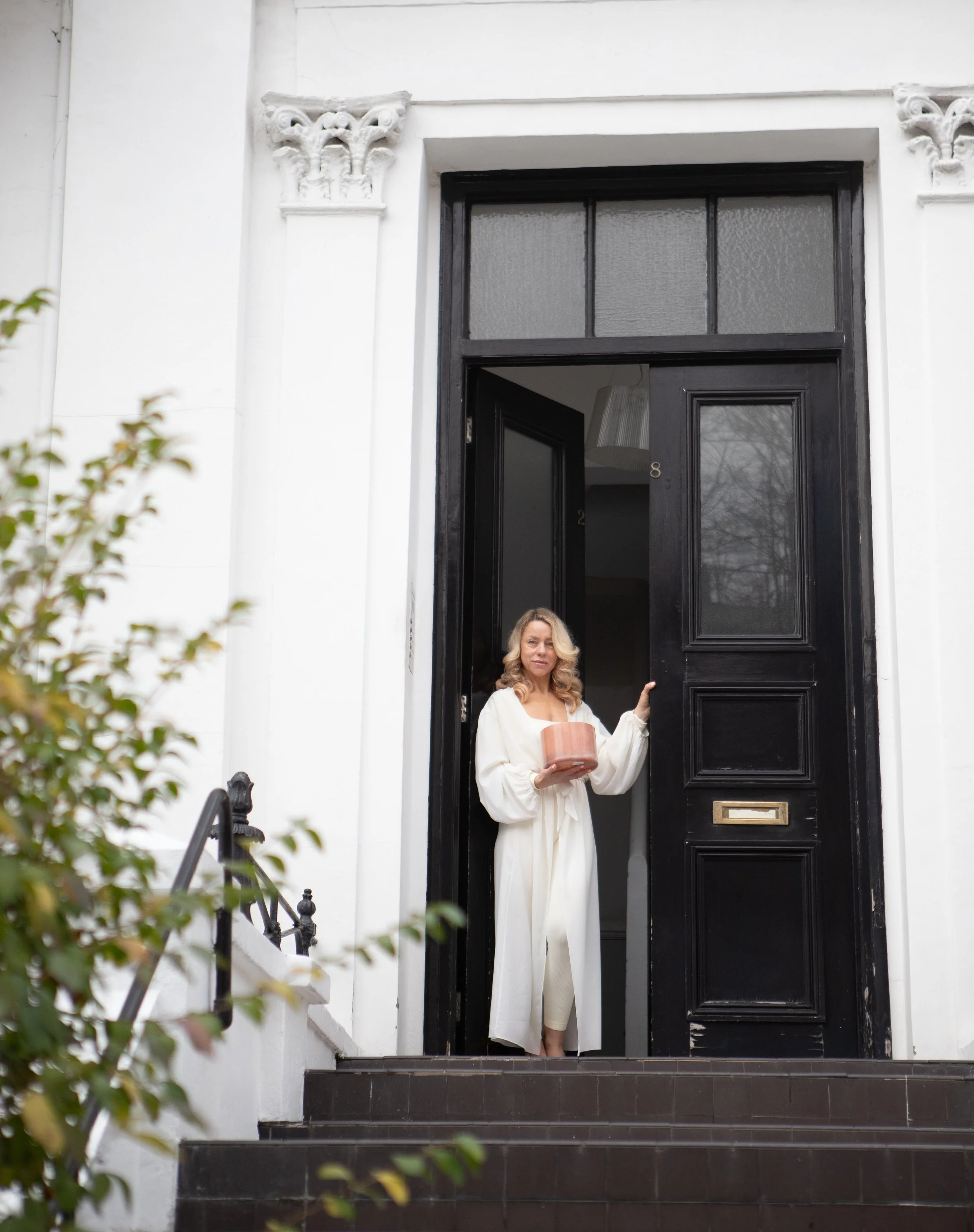 A woman with blonde hair wearing a long white robe stands on a porch, holding a pink gift box, in front of a house with a tall black door with paneling and a mail slot.