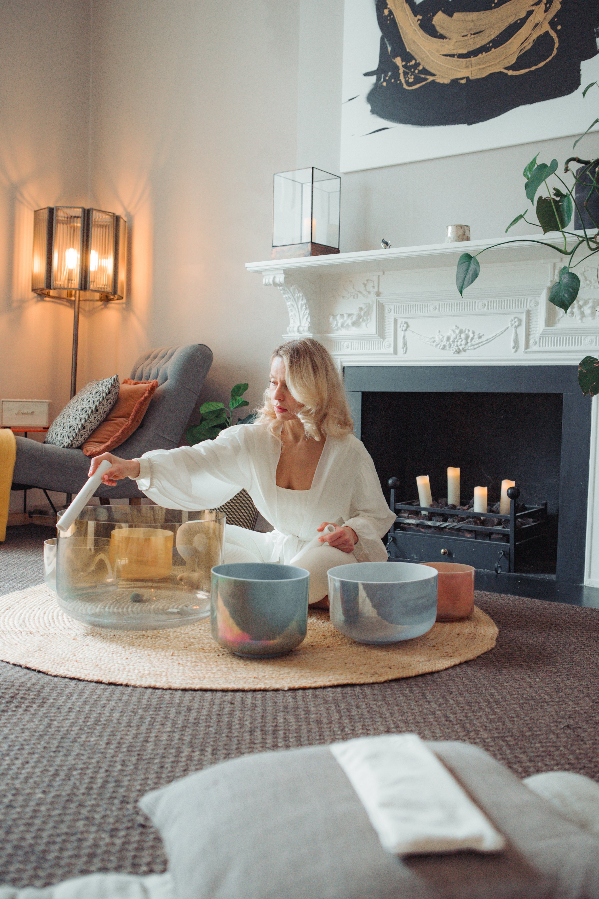 Woman sitting on a round rug in front of a fireplace, playing singing bowls, with candles on the mantle and modern decor in a cozy living room.