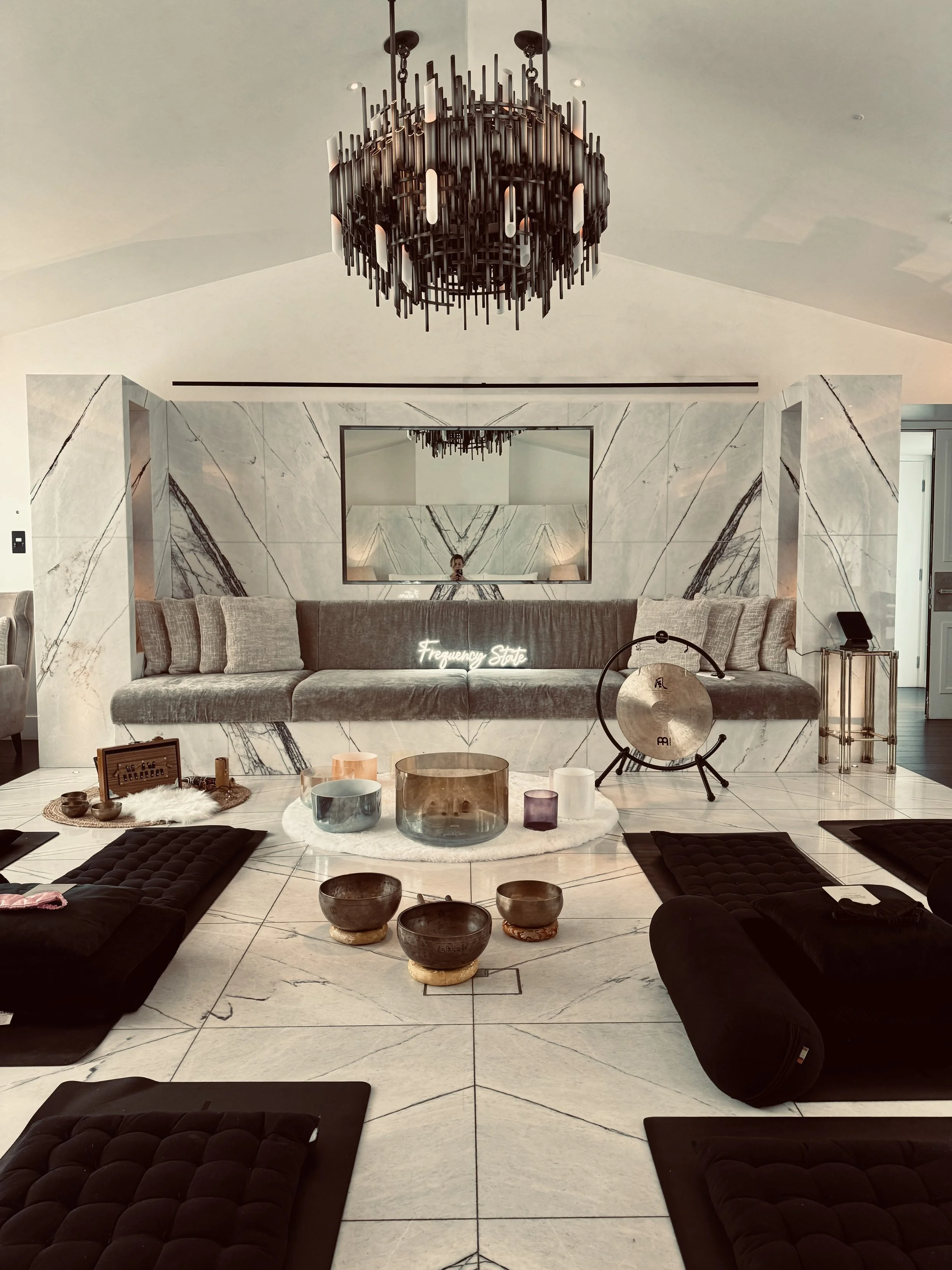 Interior of a modern living room with marble walls and floor, a large round mirror, a beige sofa with pillows, a chandelier hanging from the ceiling, and various decorative items on the coffee table and surrounding areas.