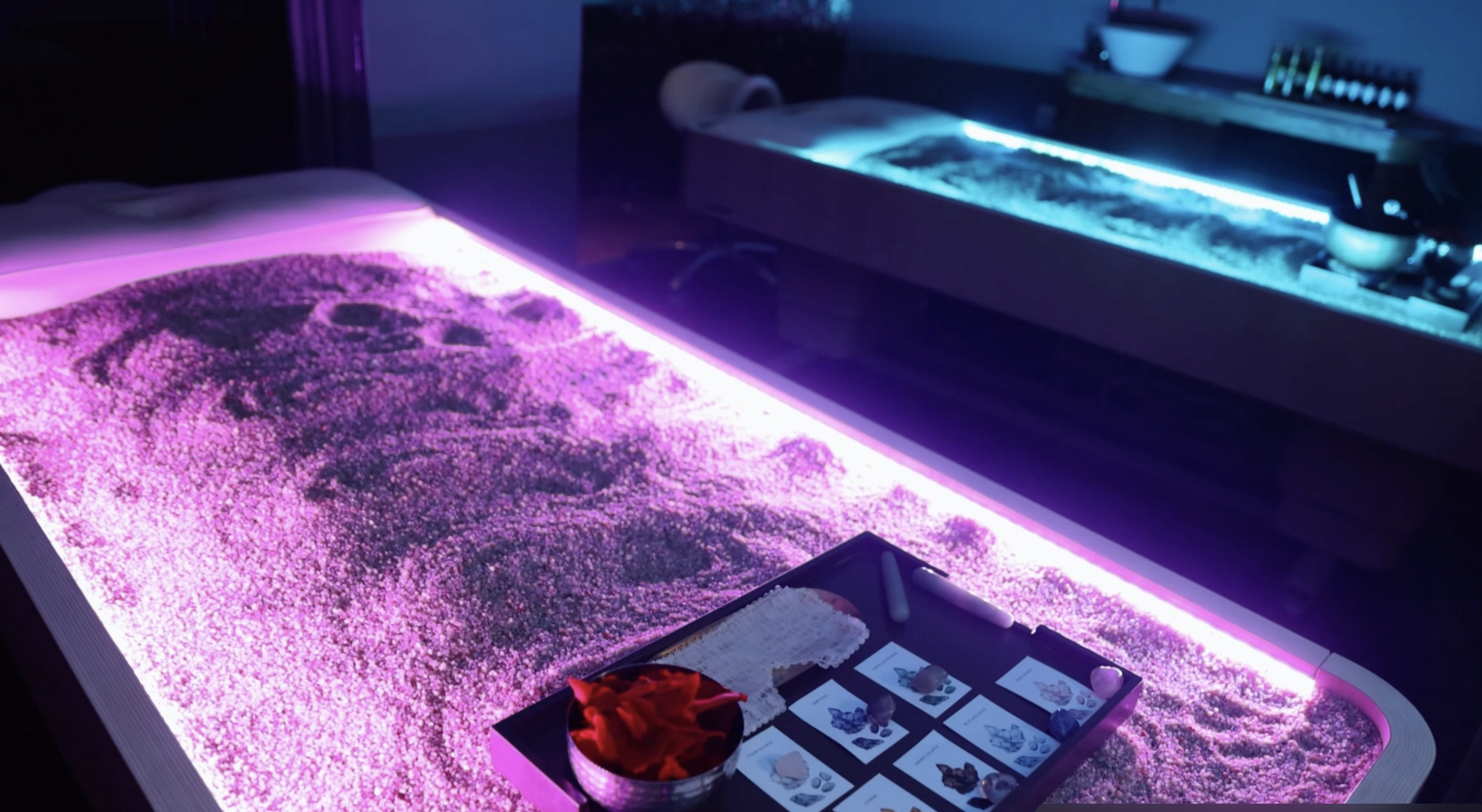 A spa room featuring two illuminated therapeutic sand beds; one under pink lighting and the other under blue lighting. A tray with massage tools and cards is placed on the pink-lit bed.